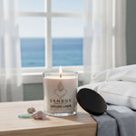A lit candle in a glass container sits on a wooden table, with a black lid and a white pillow nearby. The candle has a label that reads "SANBOX SEASIDE LINEN" and "10 OZ (2.4 US GAL)".