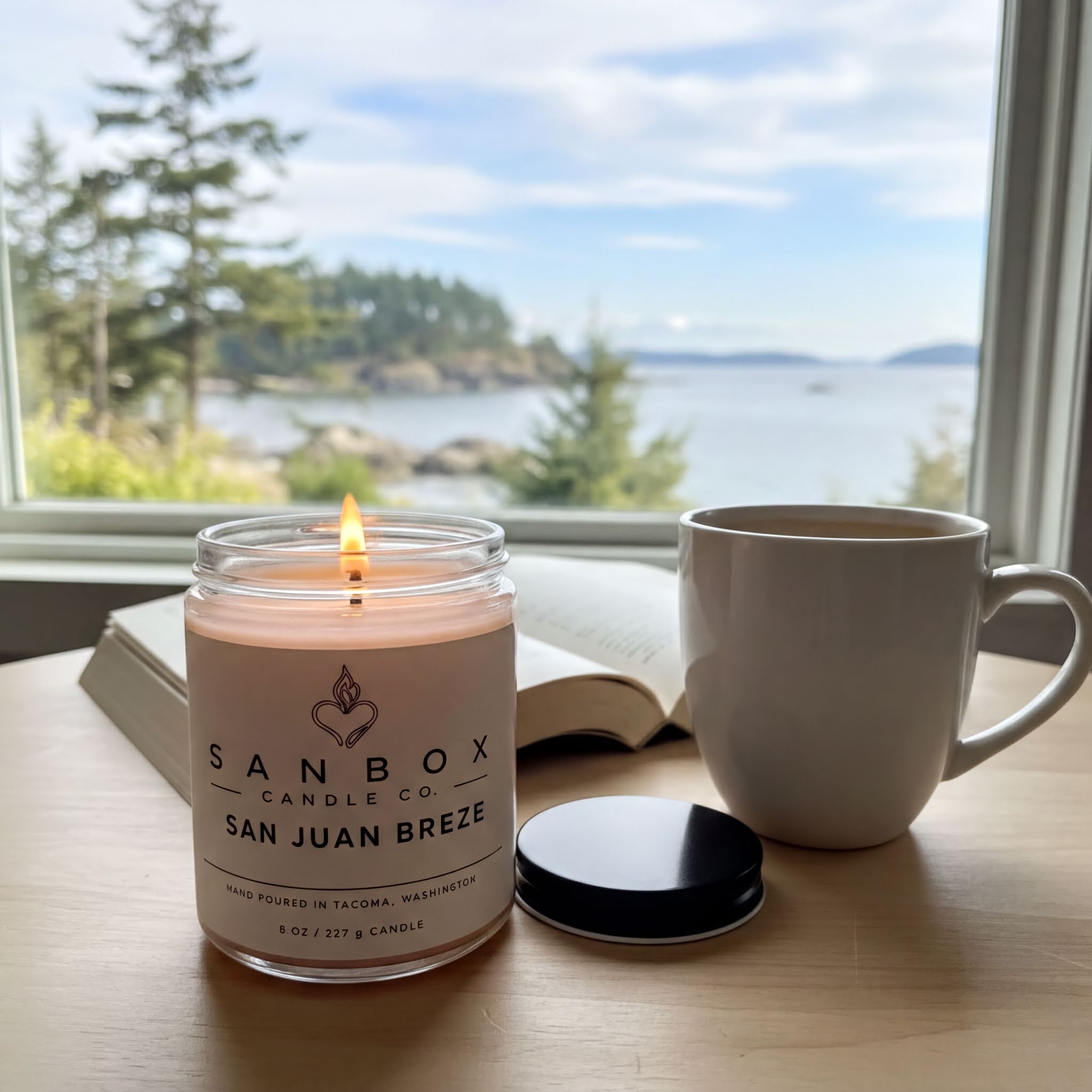 A lit candle in a jar labeled "SANBOX CO SAN JUAN BREZE" sits on a wooden table, with a white mug and an open book in the background, all overlooking a scenic view of a body of water and trees.