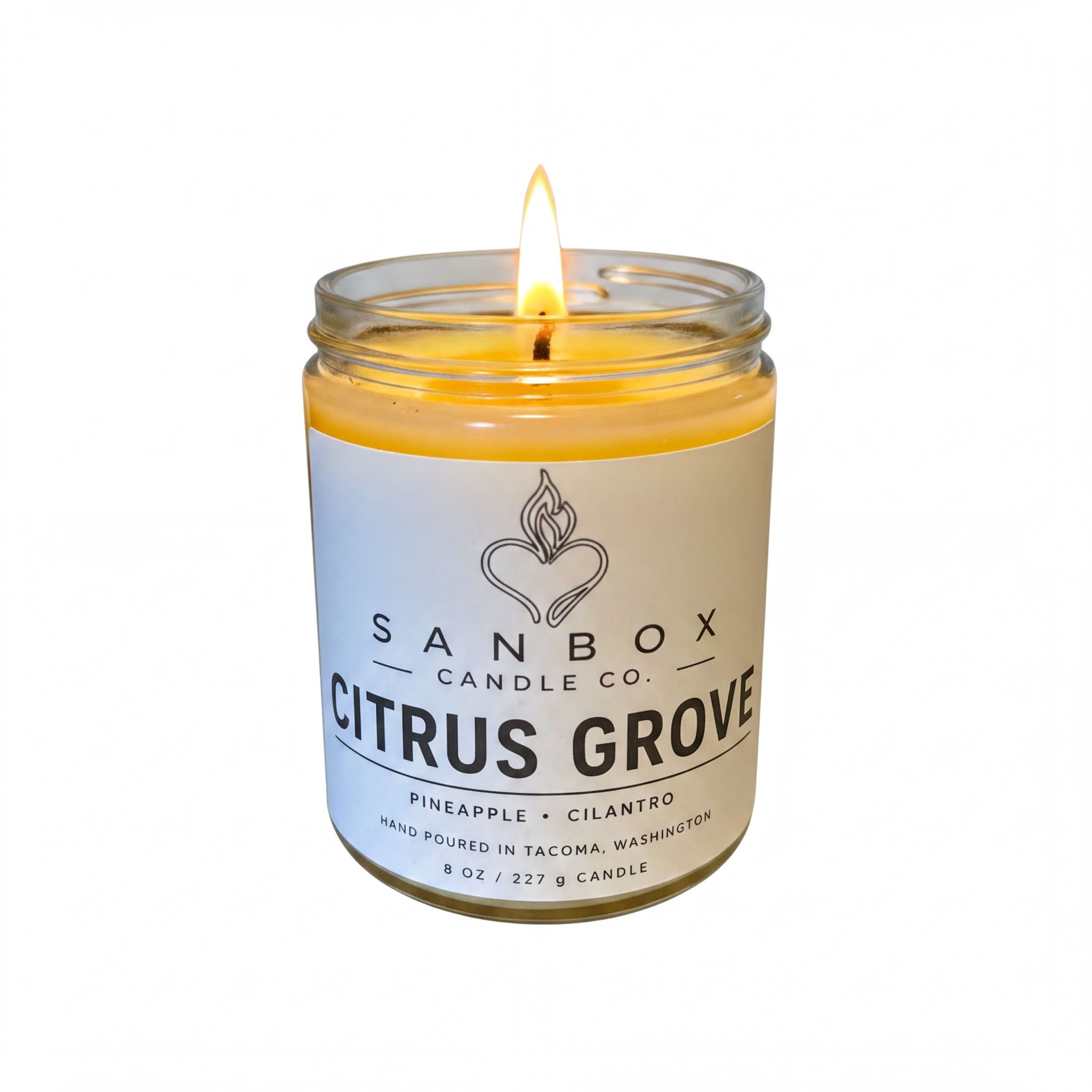 A lit candle in a glass jar with a label that reads "SANBOX CANDLE CO. CITRUS GROVE PINEAPPLE CILANTRO HAND POURED IN TACOMA WASHINGTON 8 OZ / 227g CANDLE".