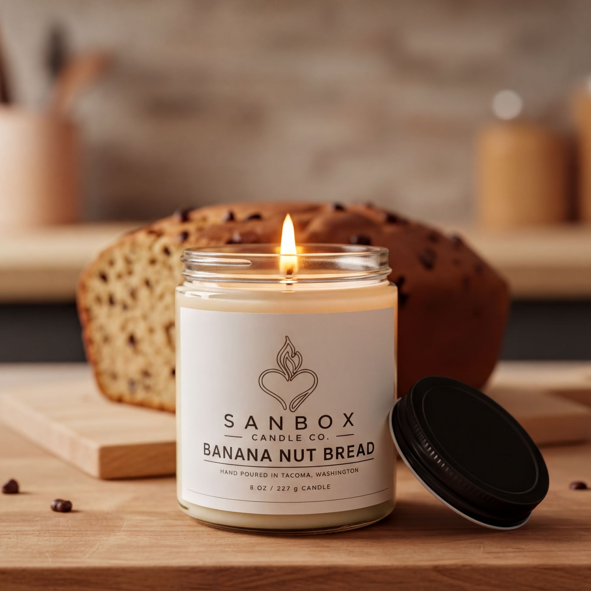 A lit candle in a glass jar with a label that reads "SANBOX CANDLE CO BANANA NUT BREAD" sits on a wooden surface, with a partially visible loaf of banana nut bread in the background.