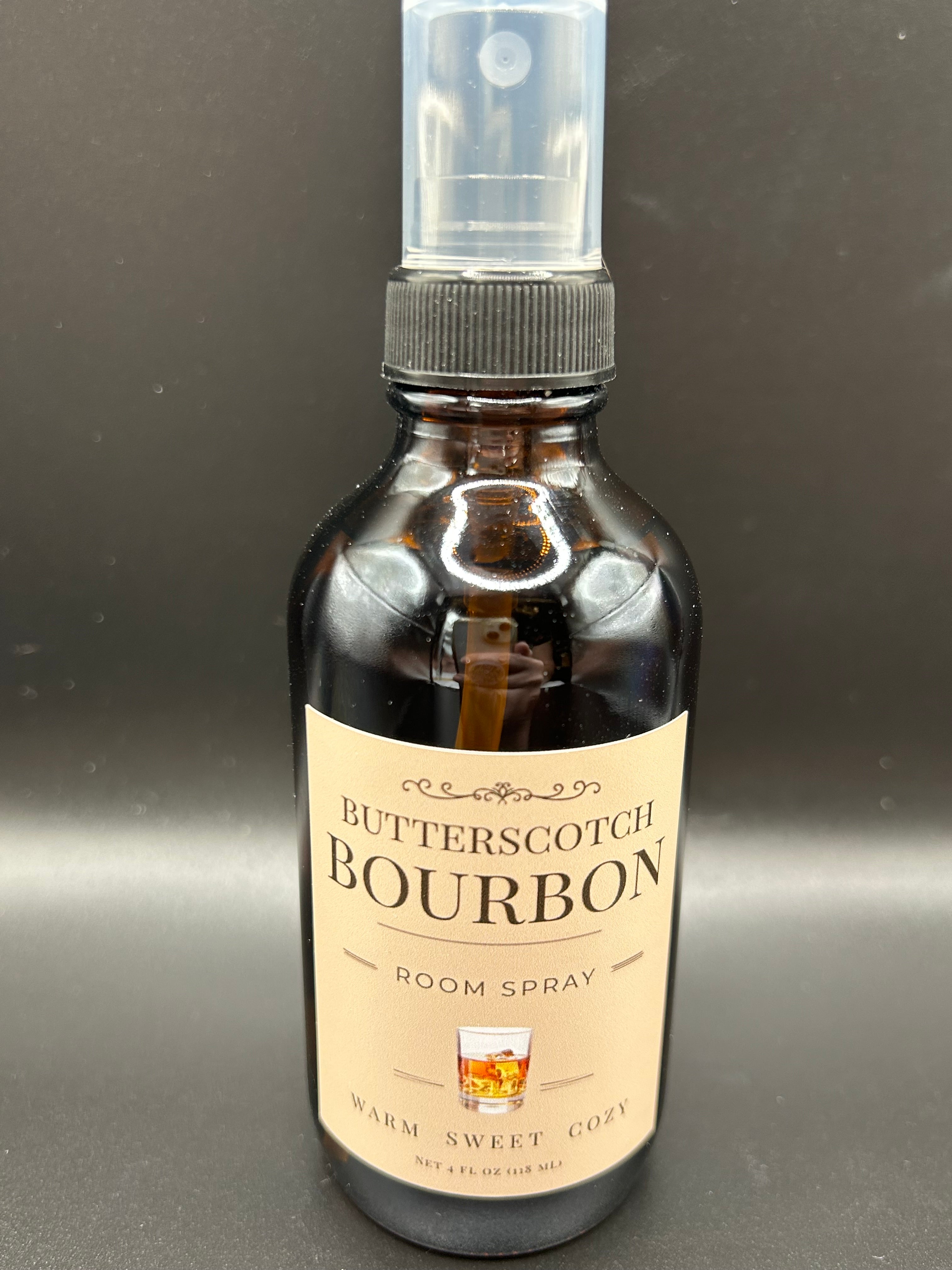 A brown glass bottle with a clear pump top, containing a room spray labeled "Butterscotch Bourbon Room Spray Warm Sweet Cozy" with a small illustration of a glass of bourbon.