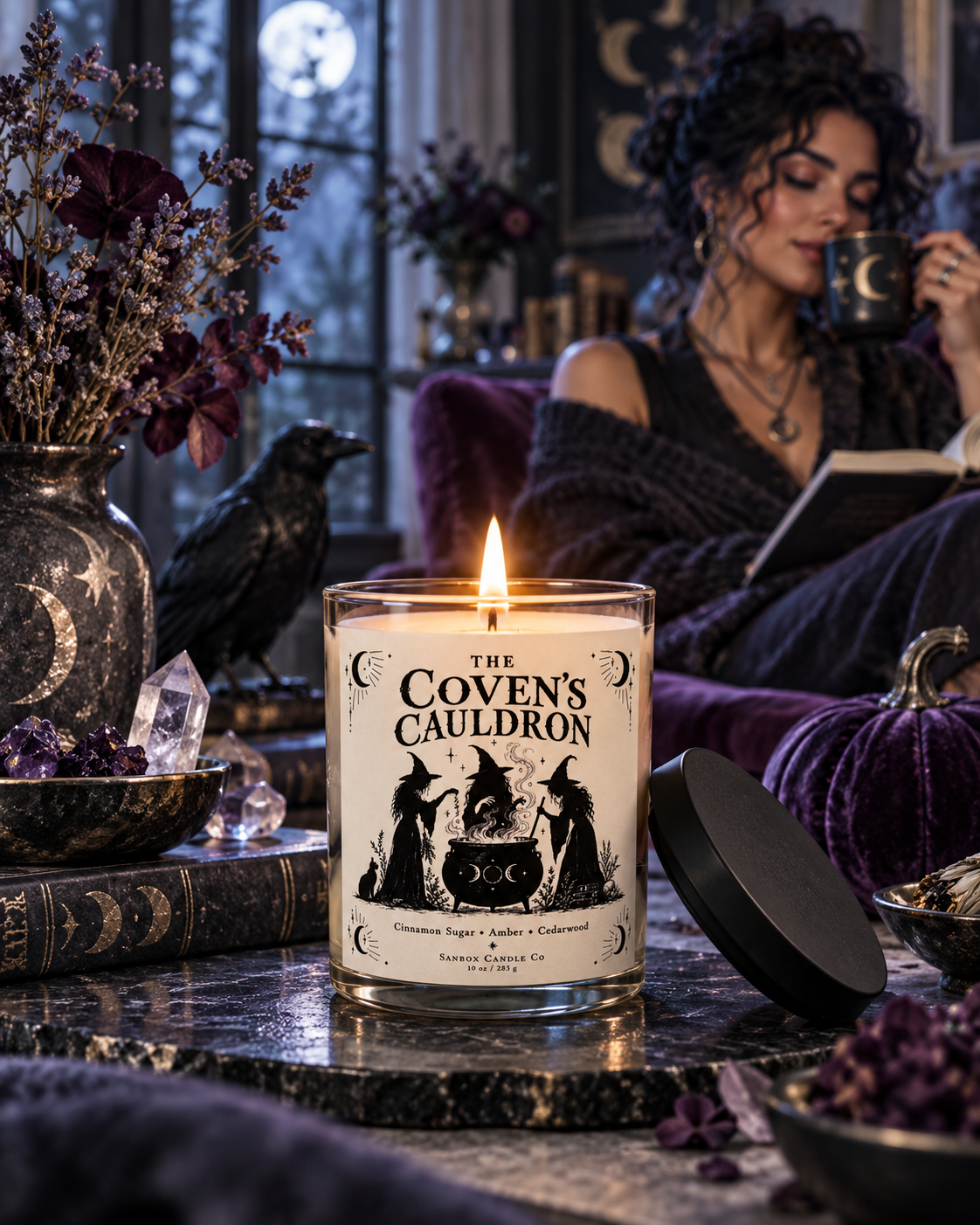 The Coven's Cauldron 10-ounce soy candle in a clear glass jar with a lit single wick and black-and-cream label showing three witches around a cauldron, styled with a black cat, raven, pumpkins, and gothic decor.