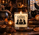 Lit 10 oz Coven's Cauldron soy candle in a clear glass jar with a cream label showing three witches around a cauldron, styled with a black cat, raven, pumpkins, autumn leaves, and gothic books.