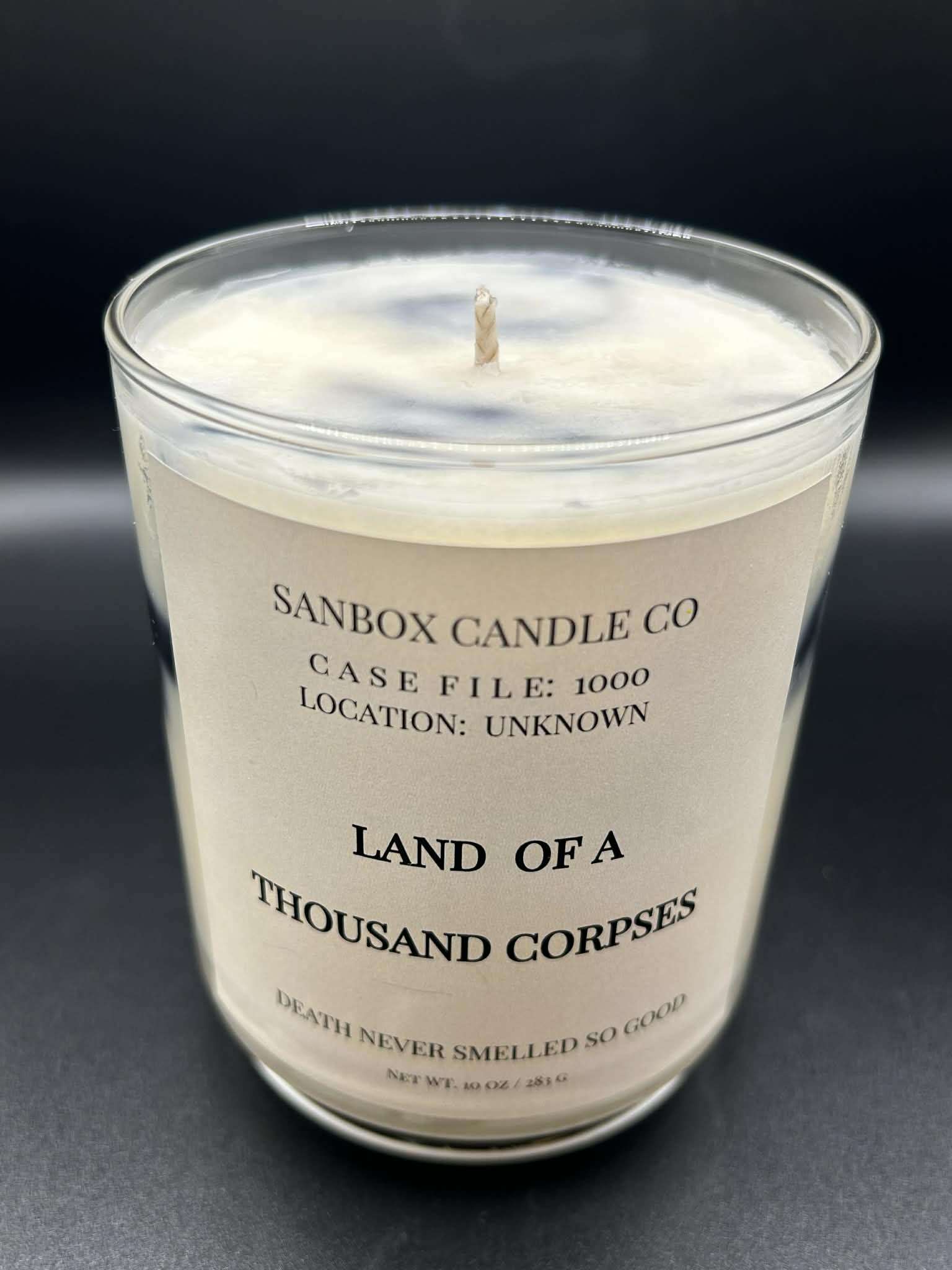 Scented candle in clear glass jar with Land of a Thousand Corpses label on black background.