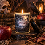Wicked Orchard 10 oz apple amber soy candle burning in a clear glass jar with a black lid, surrounded by red apples, a skull, a raven, and vintage spell books in a gothic Halloween scene