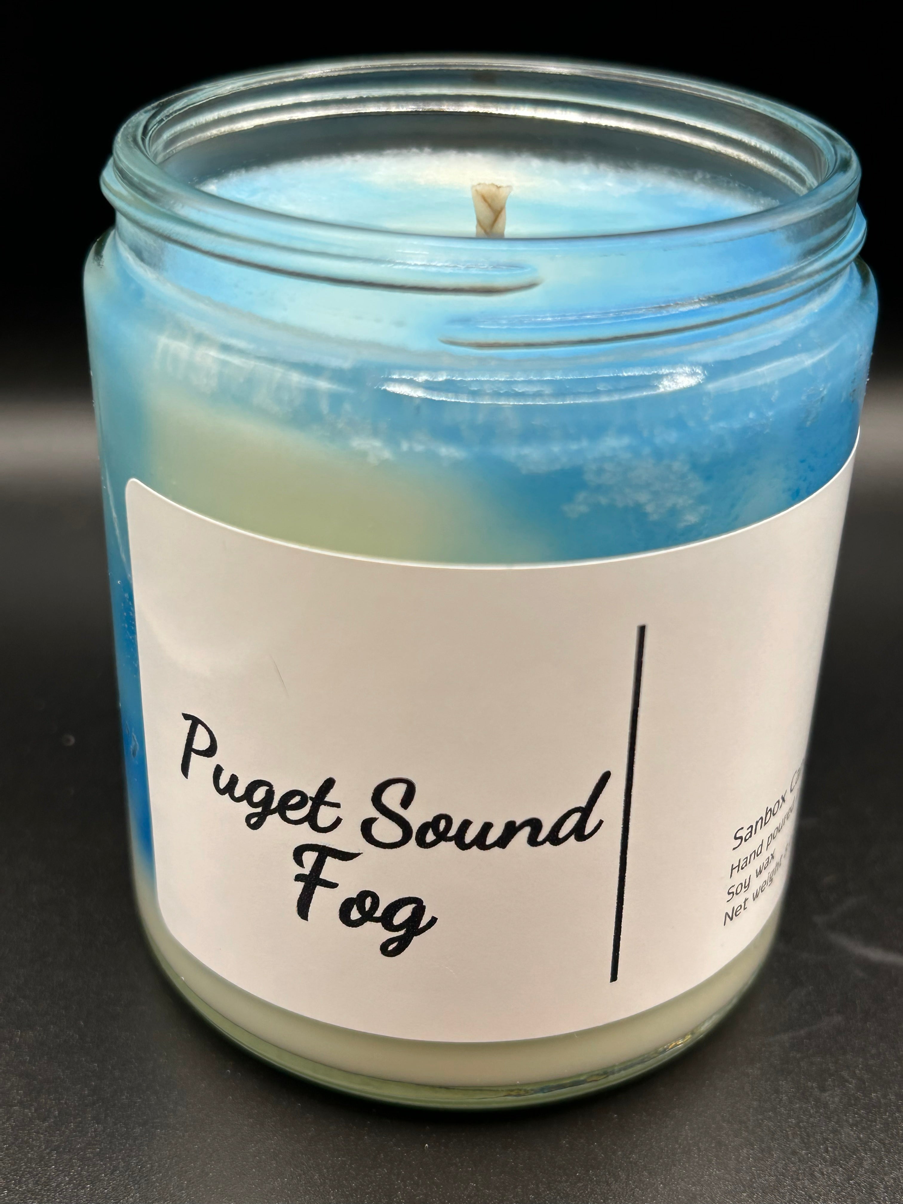 Blue and white soy candle in glass jar labeled 'Puget Sound Fog' on black background.