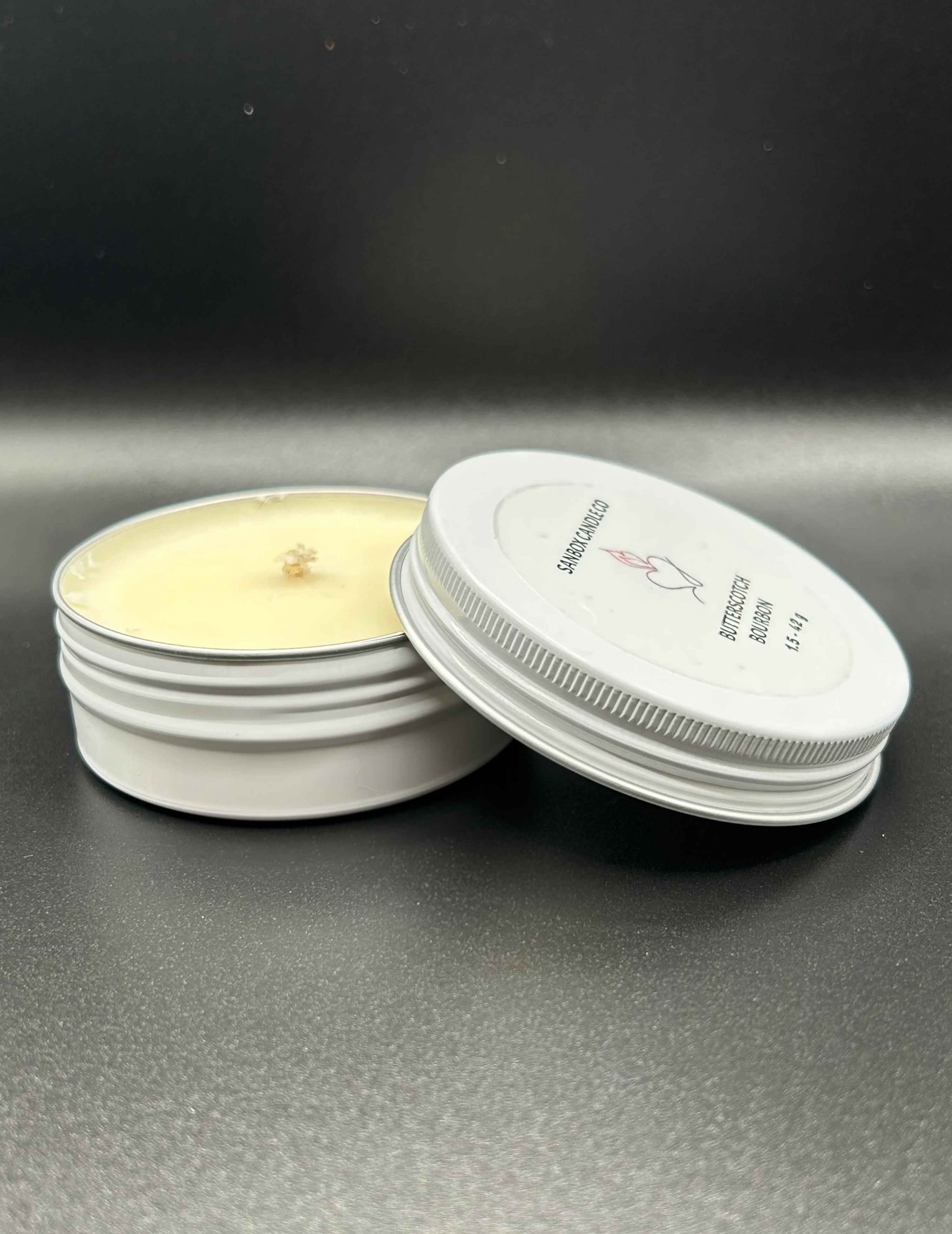 Hand-poured scented candle in a white tin with lid, displayed on a black surface.