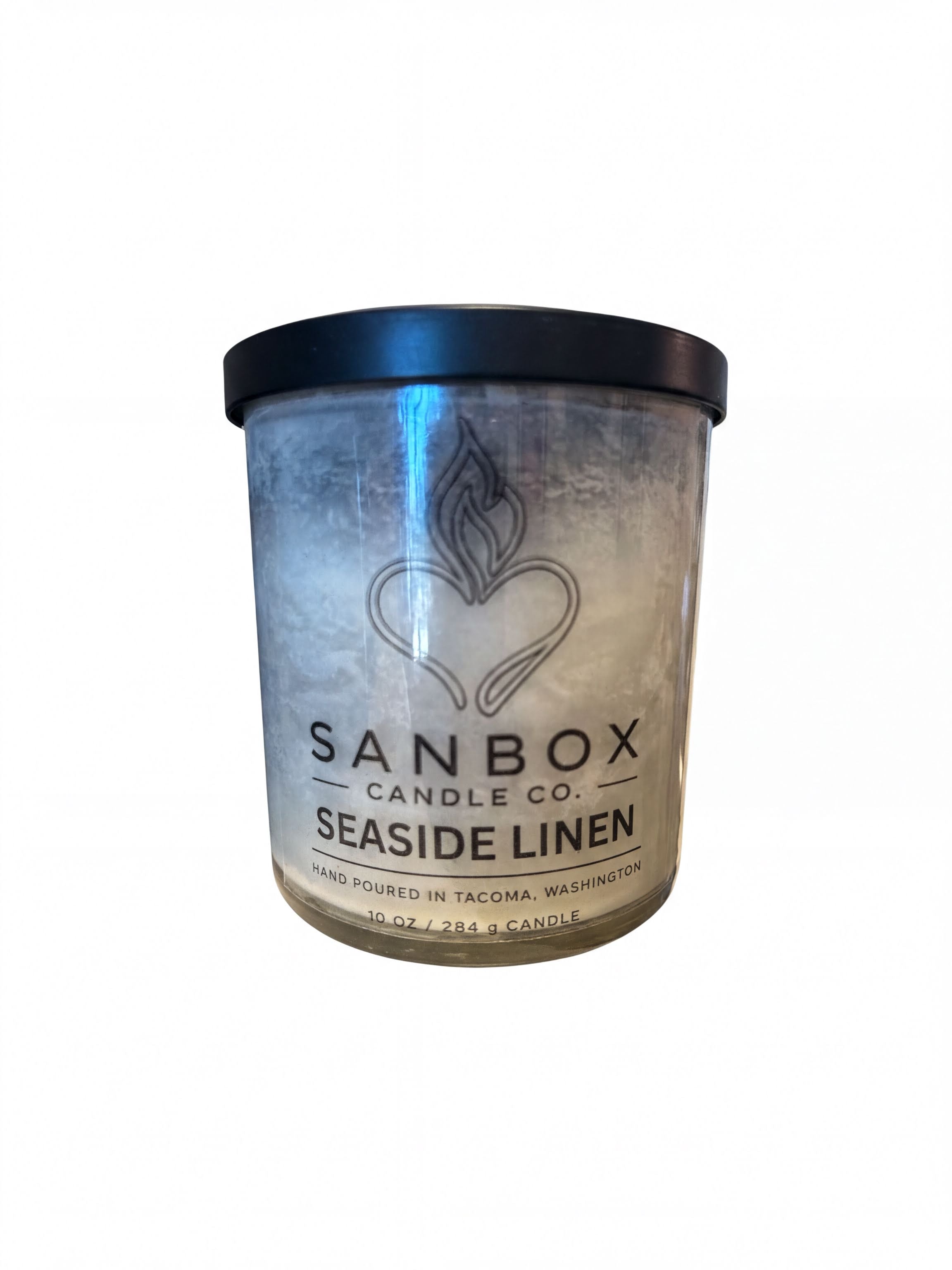 a cylindrical candle container with a black lid, labeled as a "Sanbox Candle Co. Seaside Linen" candle.