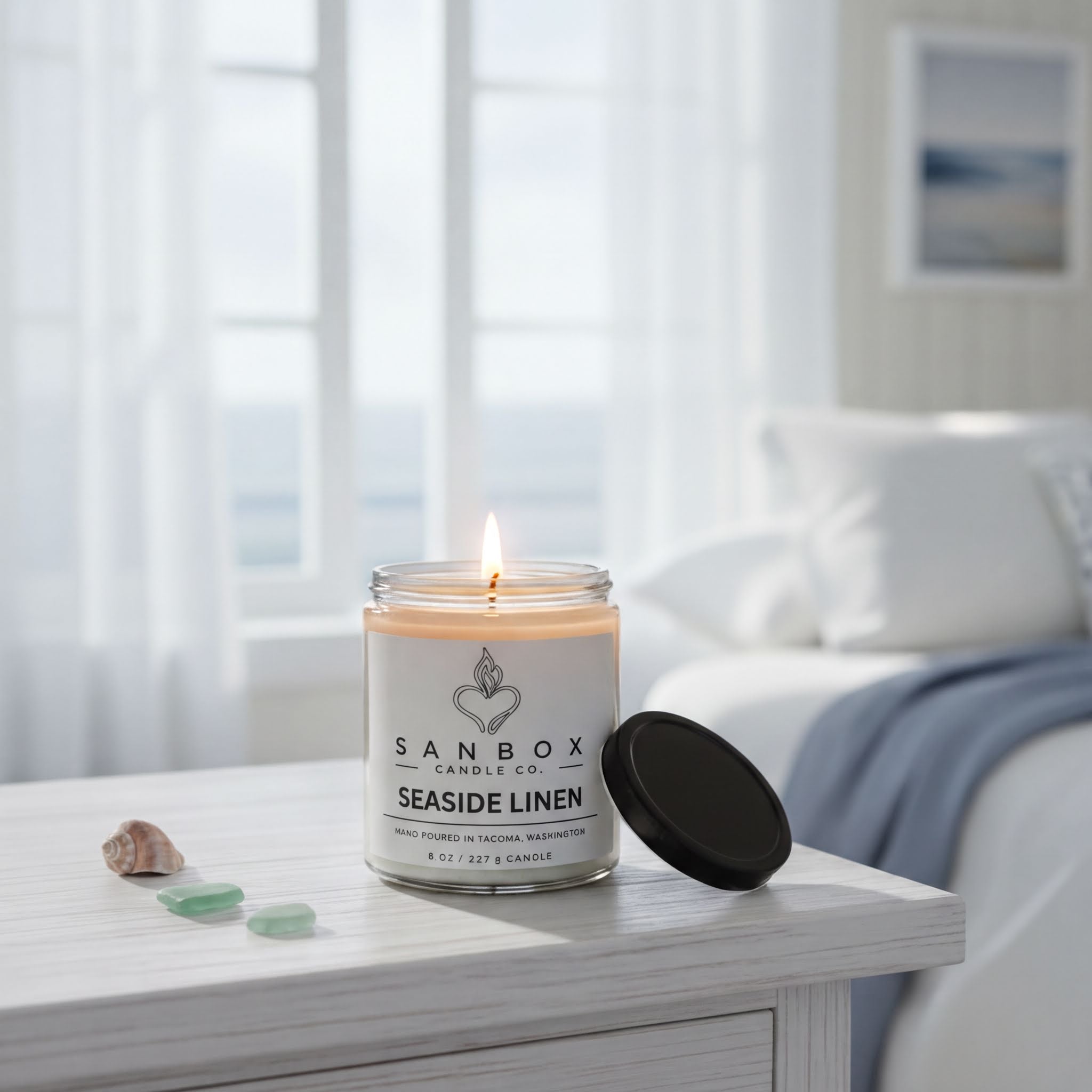 A lit candle in a jar labeled "SANBOO" sits on a white wooden table, with a black lid and a few small green stones nearby.