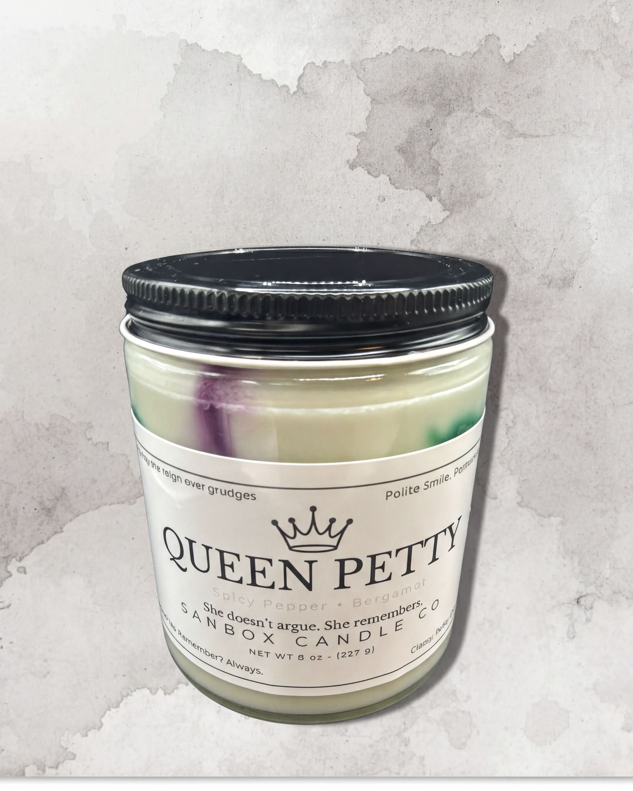 a candle labeled "Queen Petty" with a black lid, placed against a textured gray background.