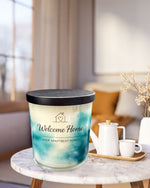 A large blue and white candle labeled "Welcome Home" sits on a marble table, accompanied by a white teapot and two white cups.