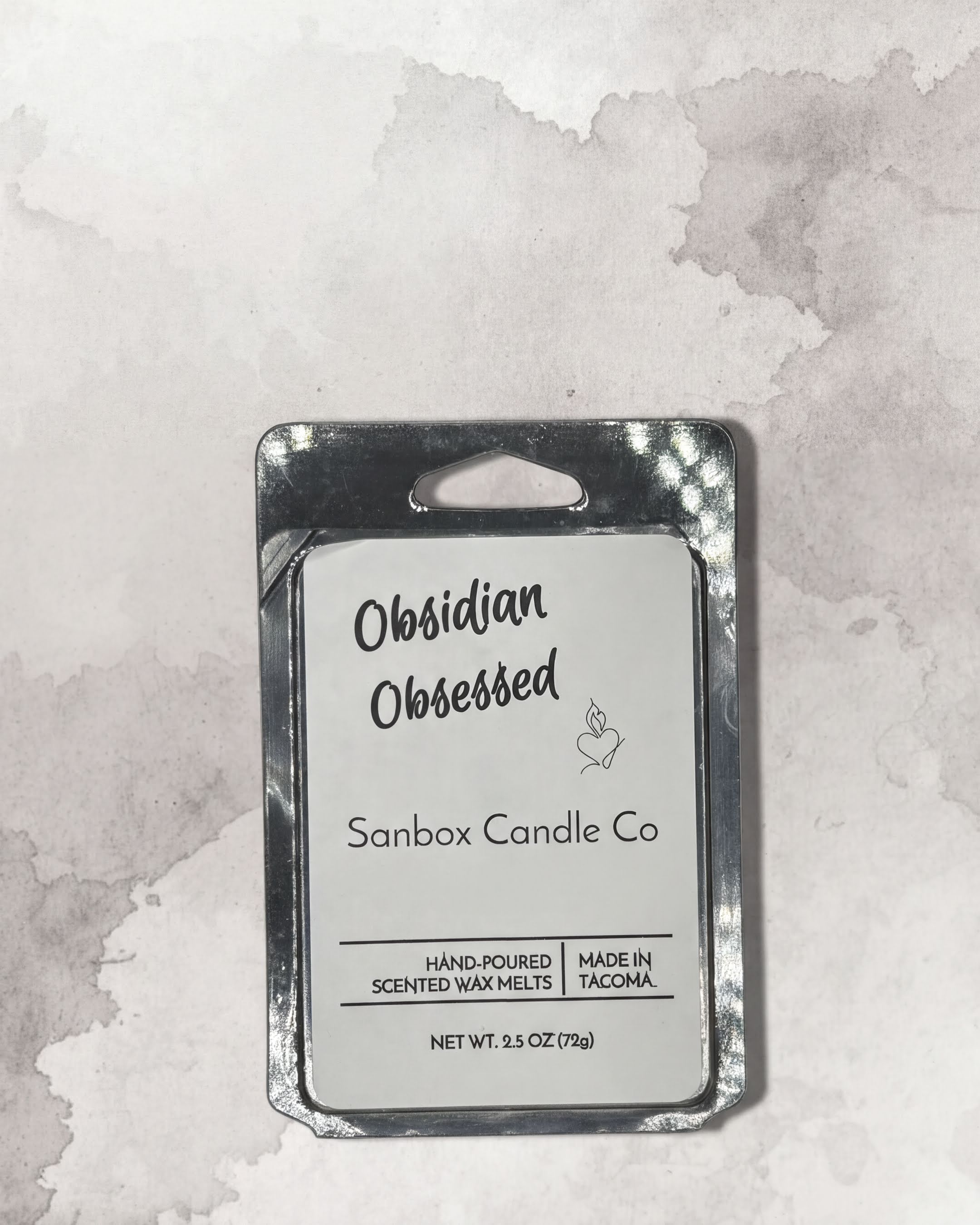 a rectangular, metallic, and silver-colored container with a label that reads "Obidian Obsessed Sanbox Candle Co."