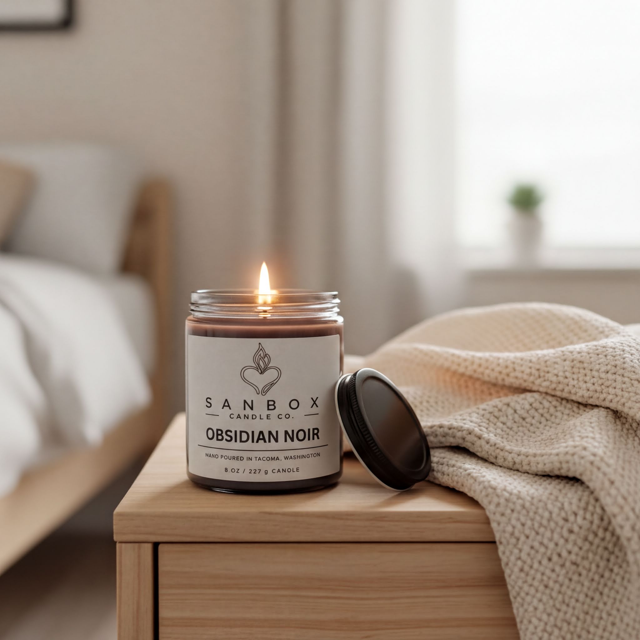 A lit candle in a jar labeled "SANBOX Candle Co. Obsidian Noir" sits on a wooden nightstand, with a beige blanket draped over it.