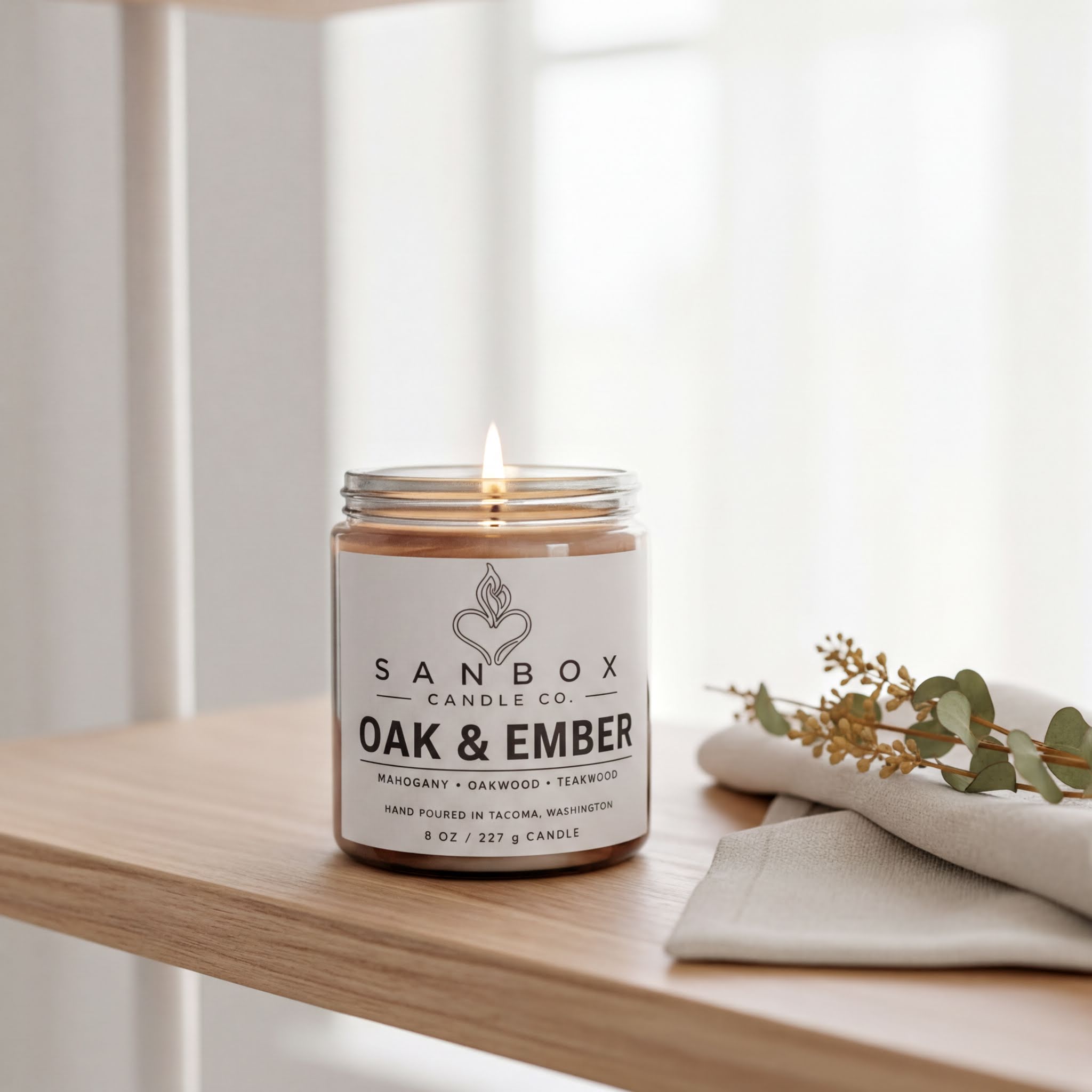 A lit candle in a jar on a wooden shelf, with a white napkin and some greenery nearby.