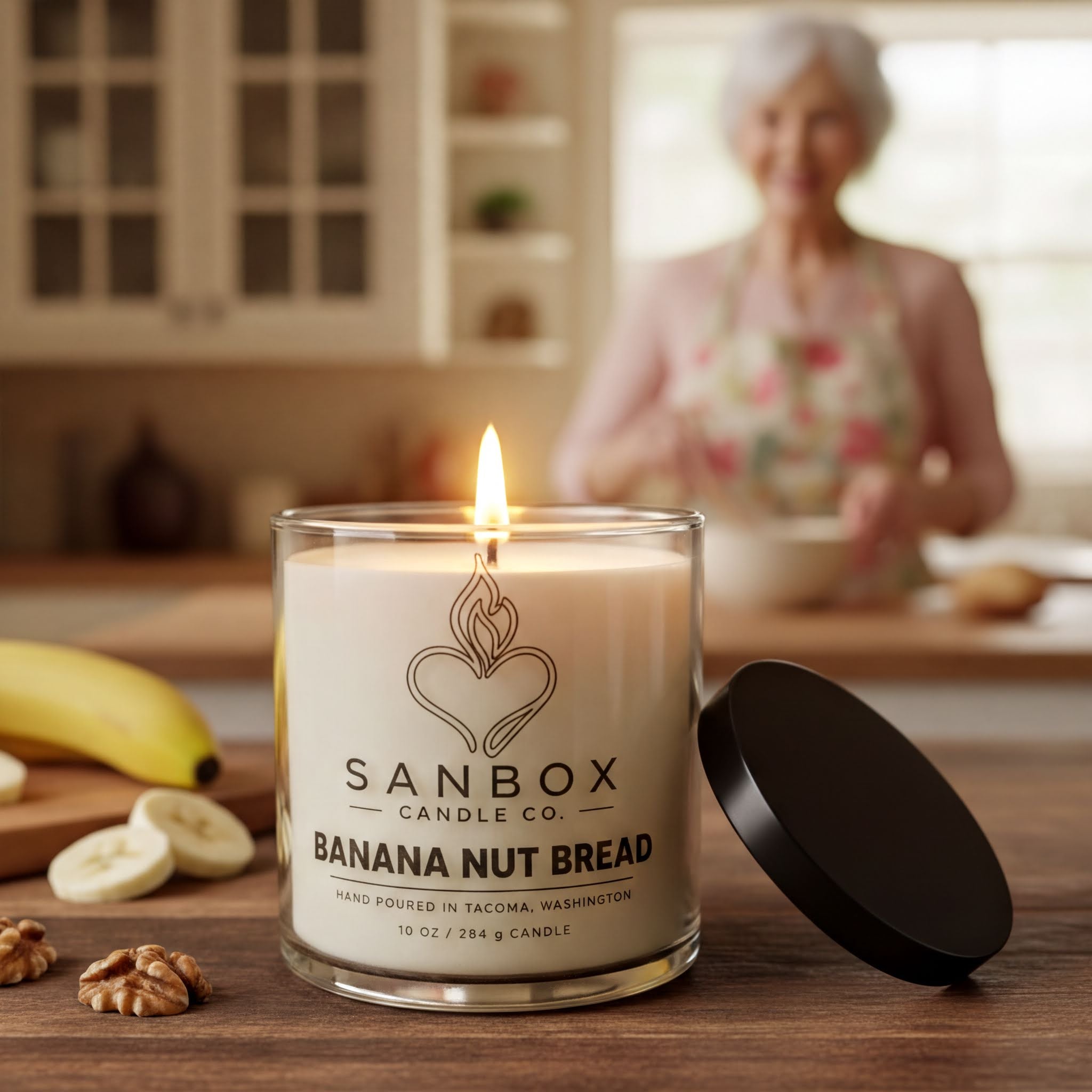 A lit candle in a glass jar labeled "SANBOX BANANA NUT BREAD" sits on a wooden table, with a blurred image of a woman in the background.