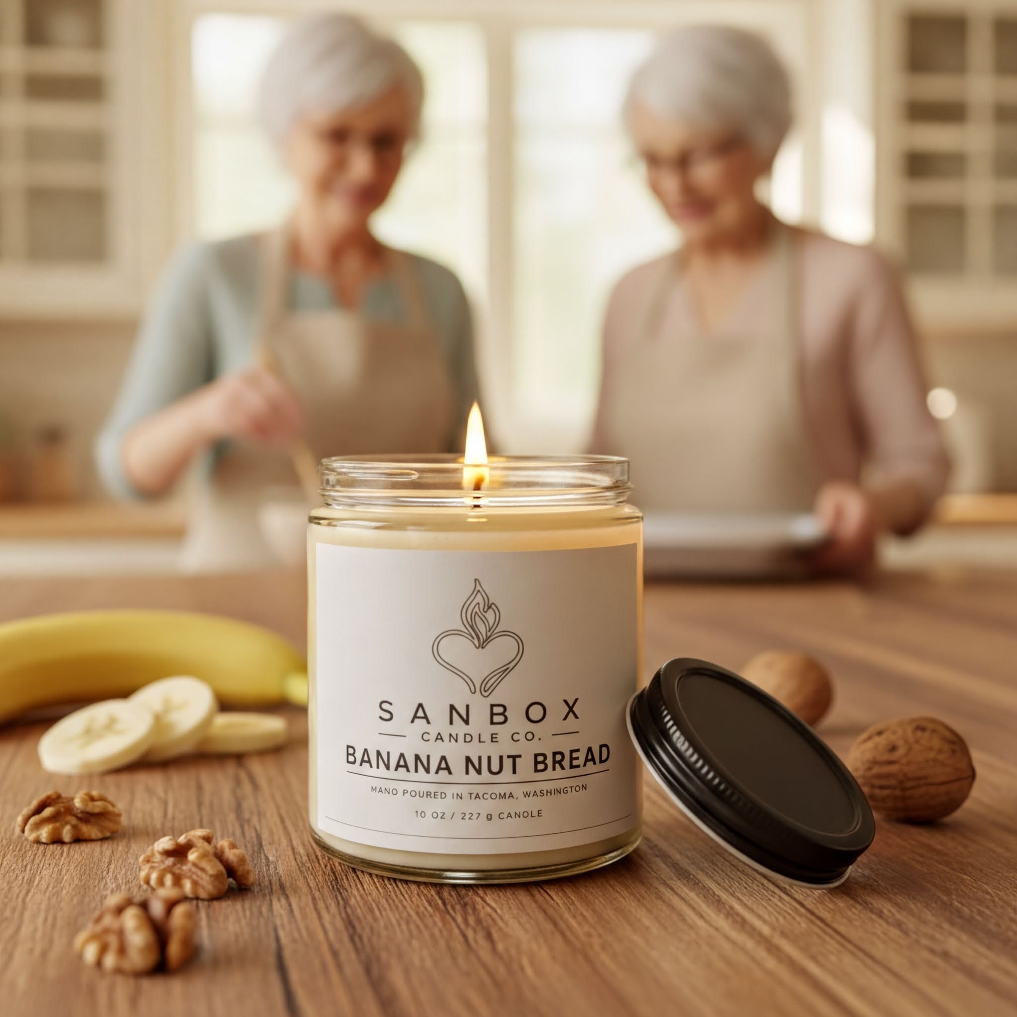 A lit candle in a glass jar labeled "SANBOX BANANA NUT BREAD" sits on a wooden table, surrounded by sliced bananas, walnuts, and a blurred background of two women.