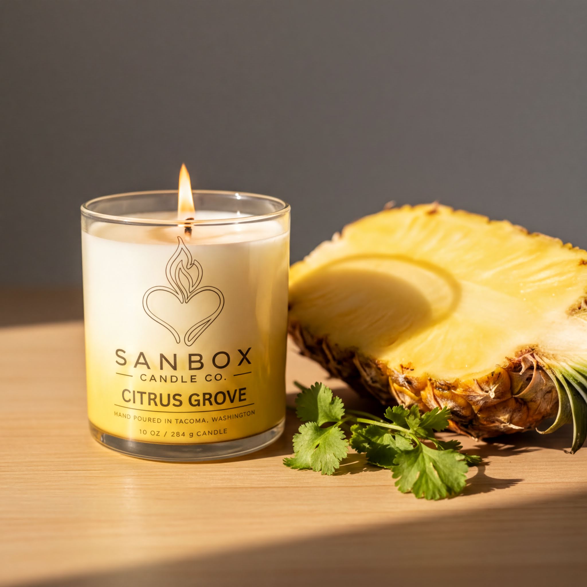 A lit candle in a glass container labeled "SANBOX CANDLE CO. CITRUS GROVE" sits next to a halved pineapple on a wooden surface.