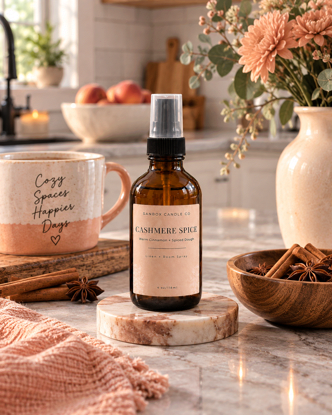 Sanbox Candle Co Cashmere Spice room spray bottle on a kitchen counter with a mug, pink flowers, cinnamon sticks, and star anise