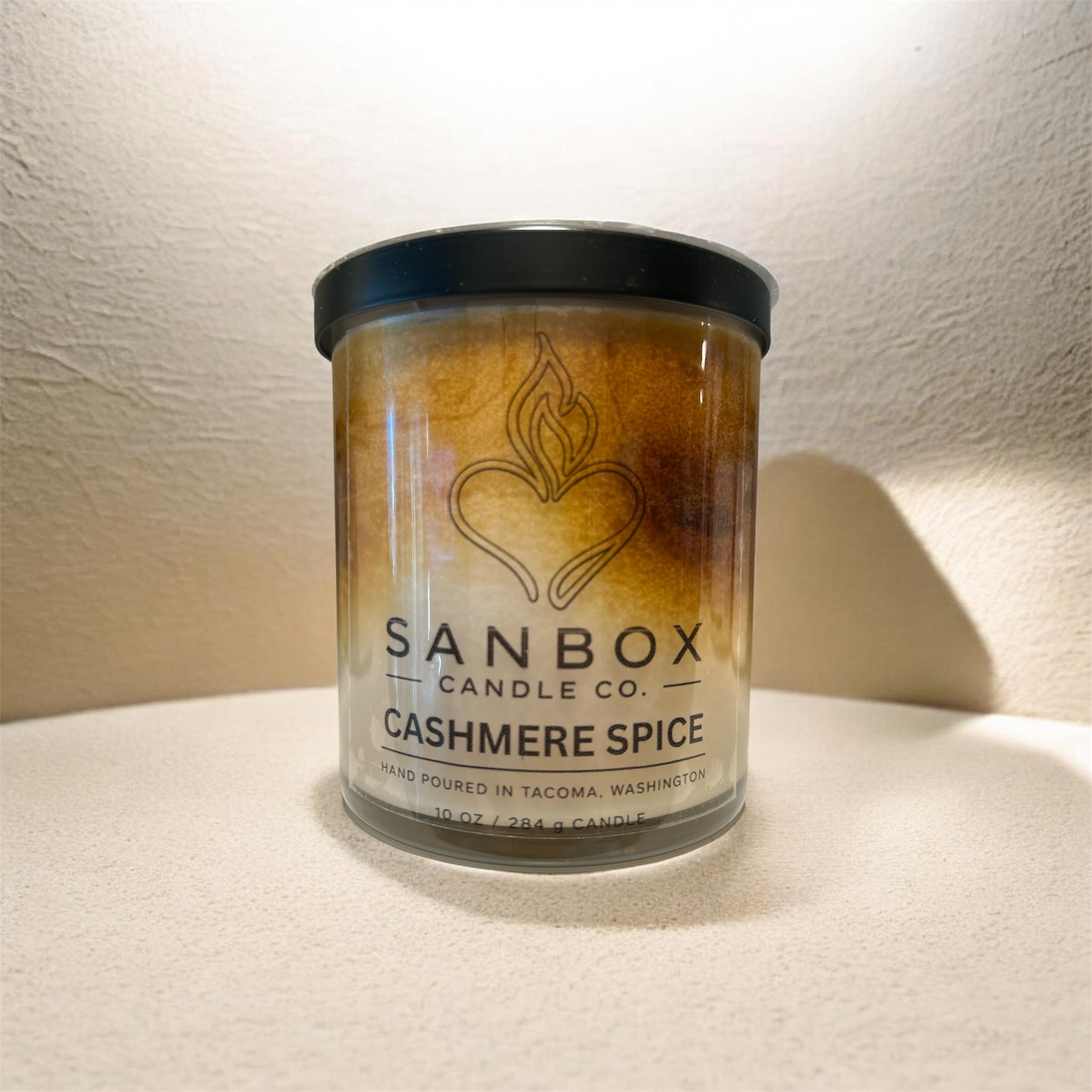 A cylindrical candle in a glass container with a label that reads "SANBOX CANDLE CO. CASHMERE SPICE".