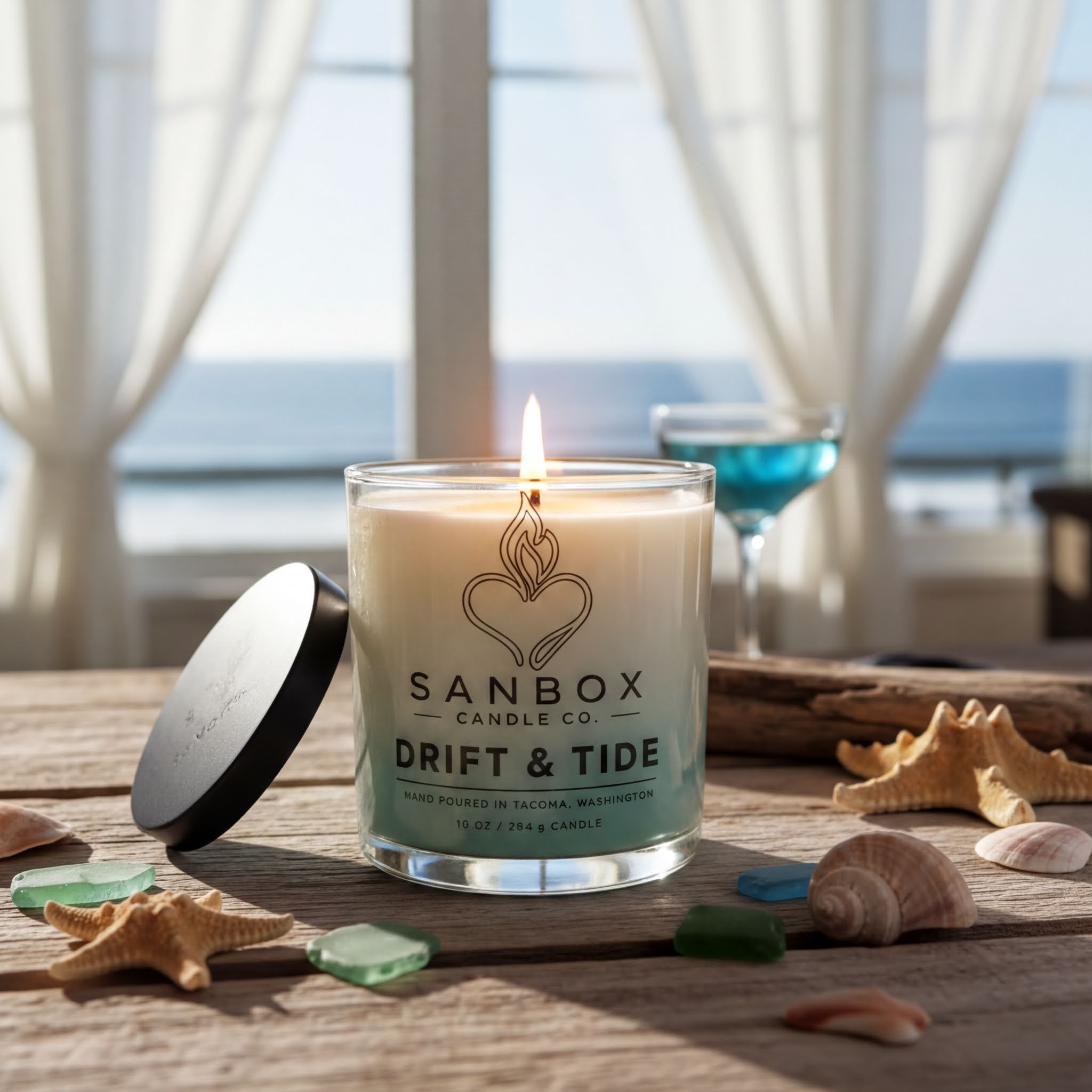 A lit candle in a glass jar labeled "Drift & Tide" sits on a wooden table, surrounded by seashells and a glass of blue liquid, with a view of the ocean through a window in the background.