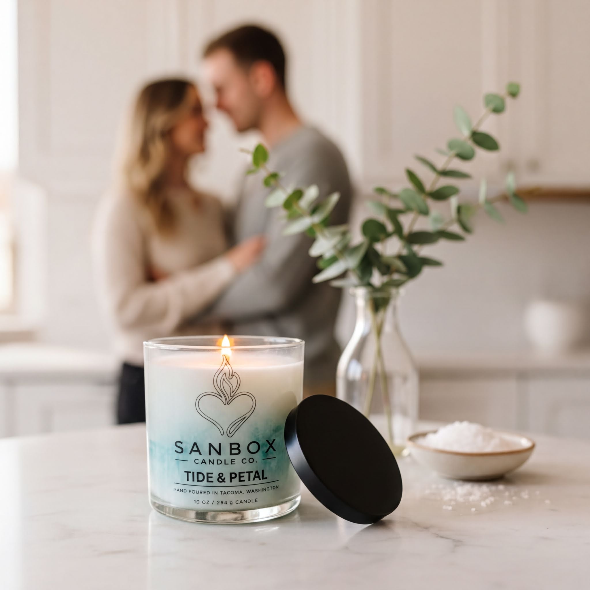a candle labeled "Sanbox Tide & Petal" on a marble countertop, with a blurred couple in the background.