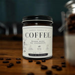 A jar of Sanbox Candle Co. Coffee candle with a black lid and white label, placed on a wooden surface with coffee beans scattered around it.
