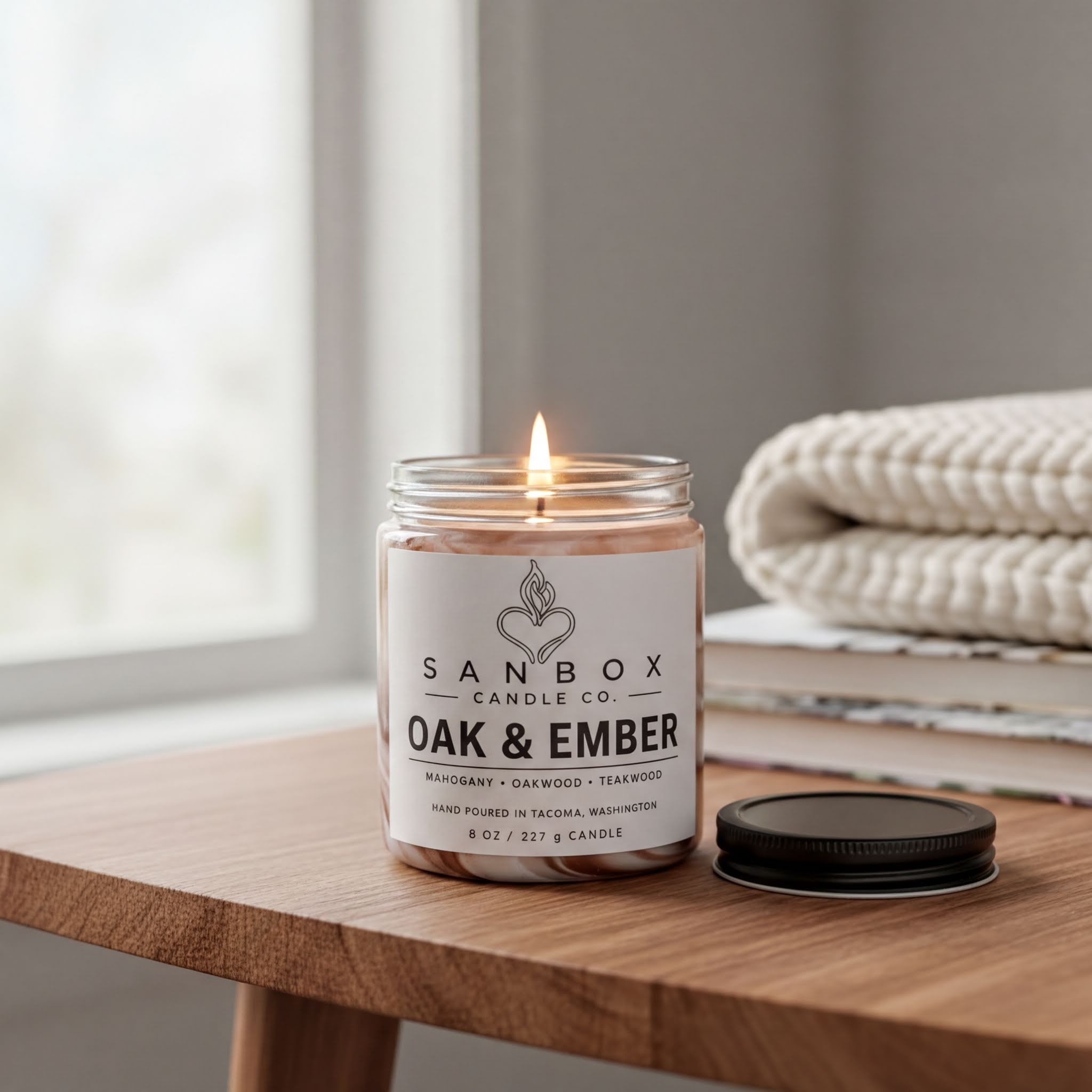 A lit candle in a glass jar on a wooden table, with a white towel and a stack of books in the background.