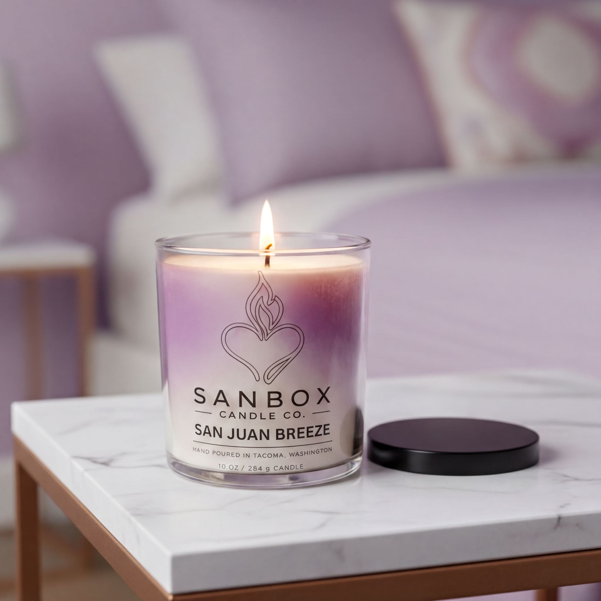 A lit lavender-colored candle in a glass jar with a black lid sits on a white marble table, with a purple pillow visible in the background.