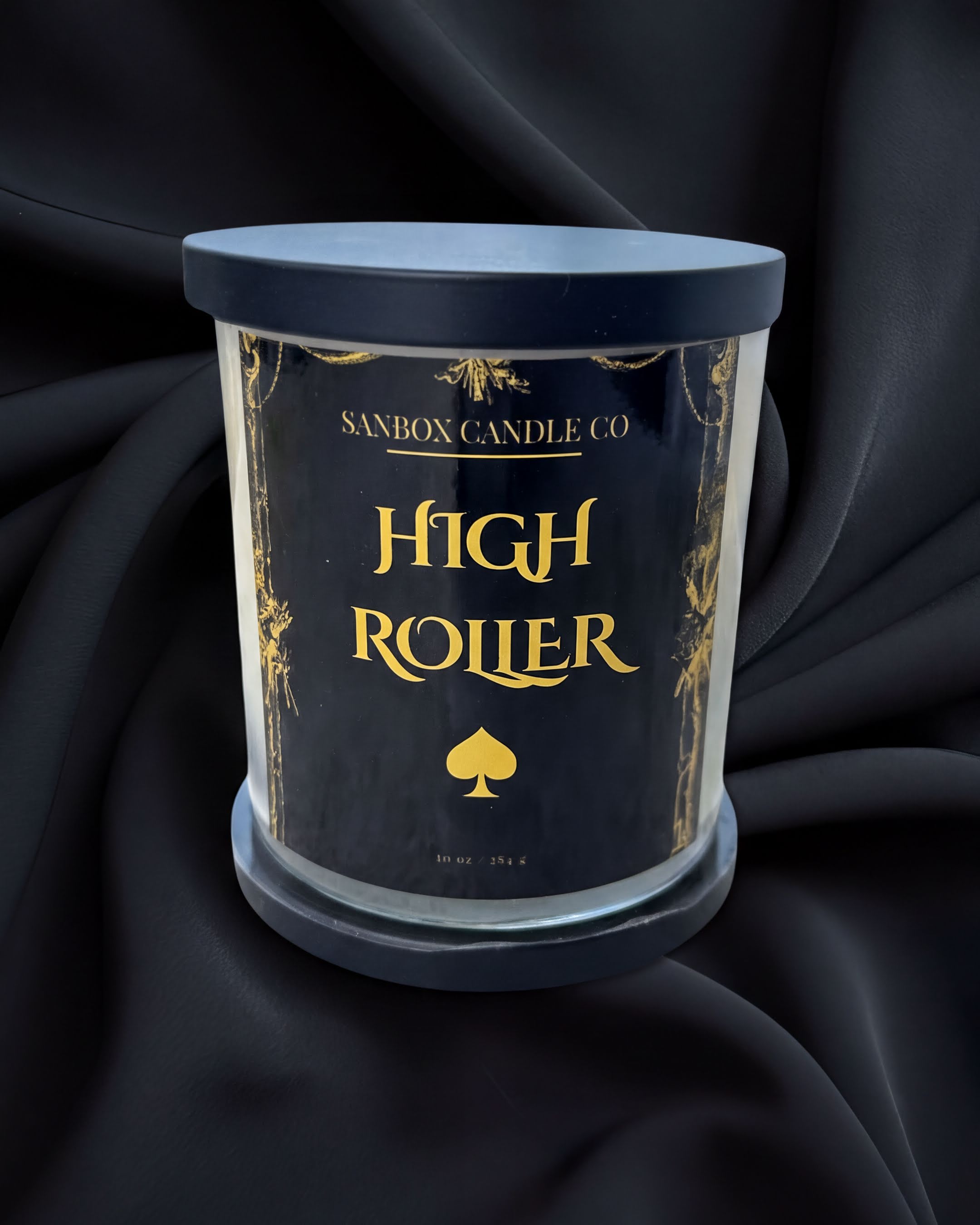 a candle labeled "High Roller" in a black and gold color scheme, placed on a black surface against a dark background.