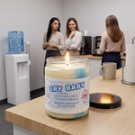 a candle labeled " Cry Baby" on a wooden table, with two women standing in the background.