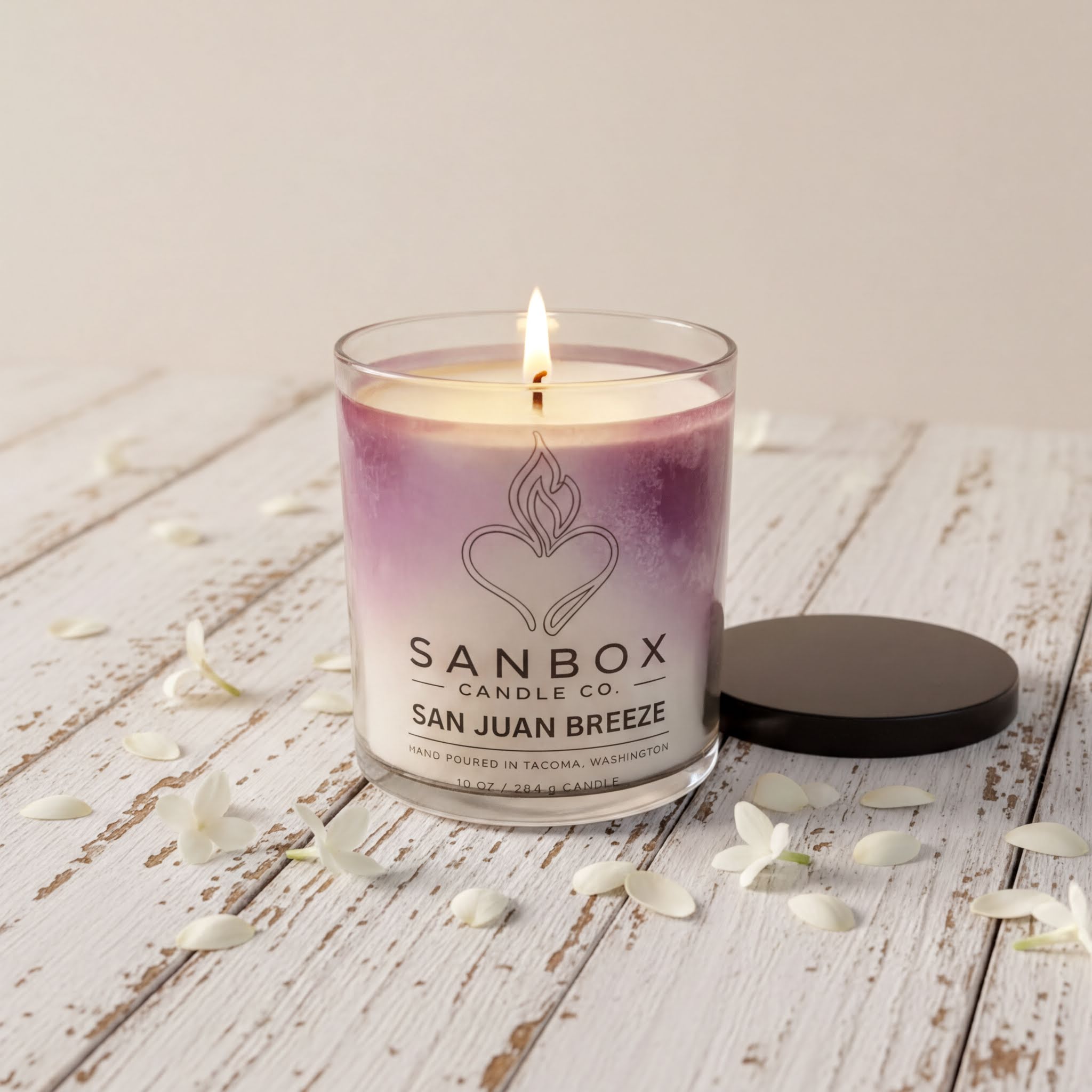 A lit candle in a glass container with a purple and white label, placed on a wooden surface with white flower petals around it.