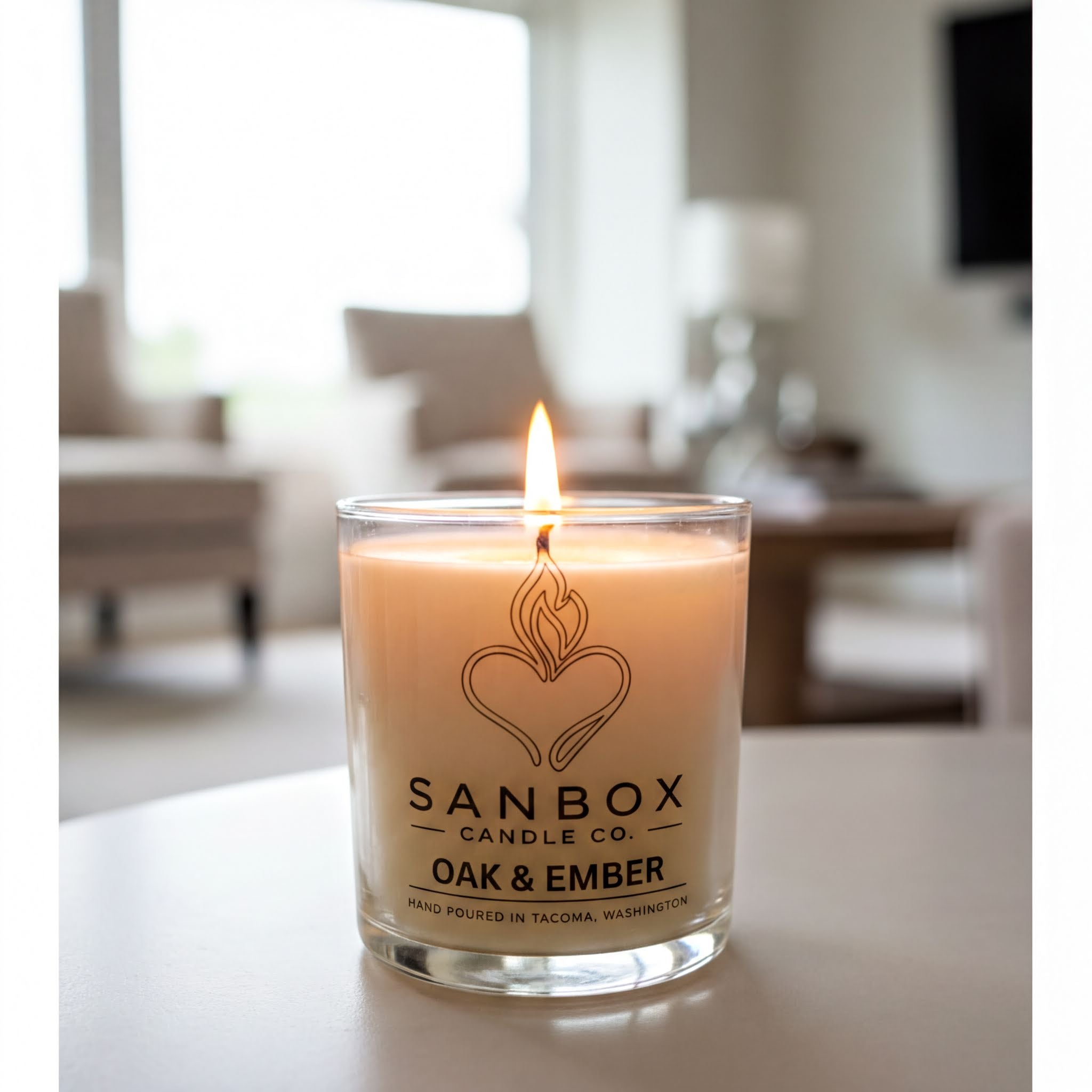 A lit candle in a glass container with a label that reads "SANBOX CANDLE CO. OAK & EMBER" and "HAND POULED IN TACOMA, WASHINGTON".