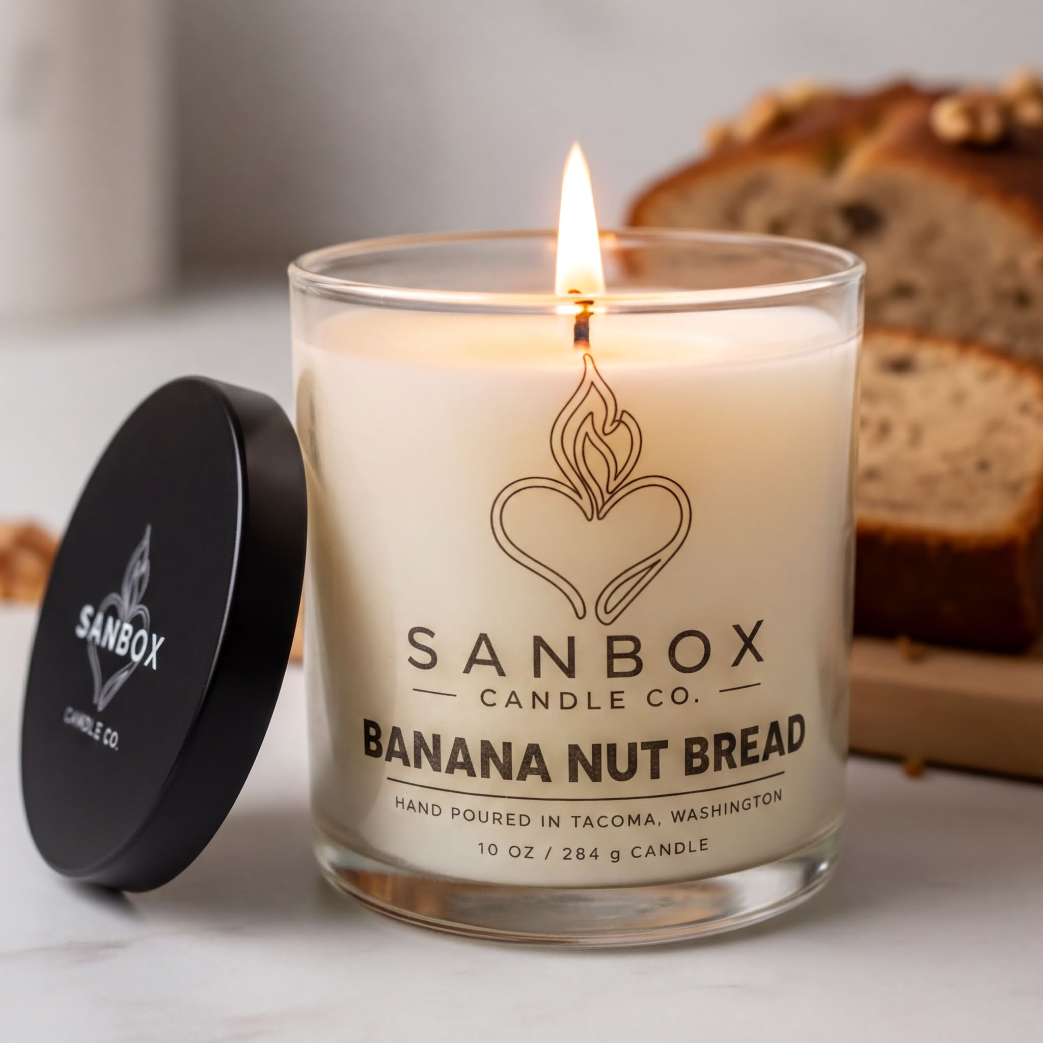 A lit candle in a glass container with a label that reads "SANBOX CANDLE CO. BANANA NUT BREAD" and "HAND POURED IN TACOMA, WASHINGTON 10 OZ / 286 Q TICKET". The candle is placed next to a black lid and a sliced loaf of banana nut bread.
