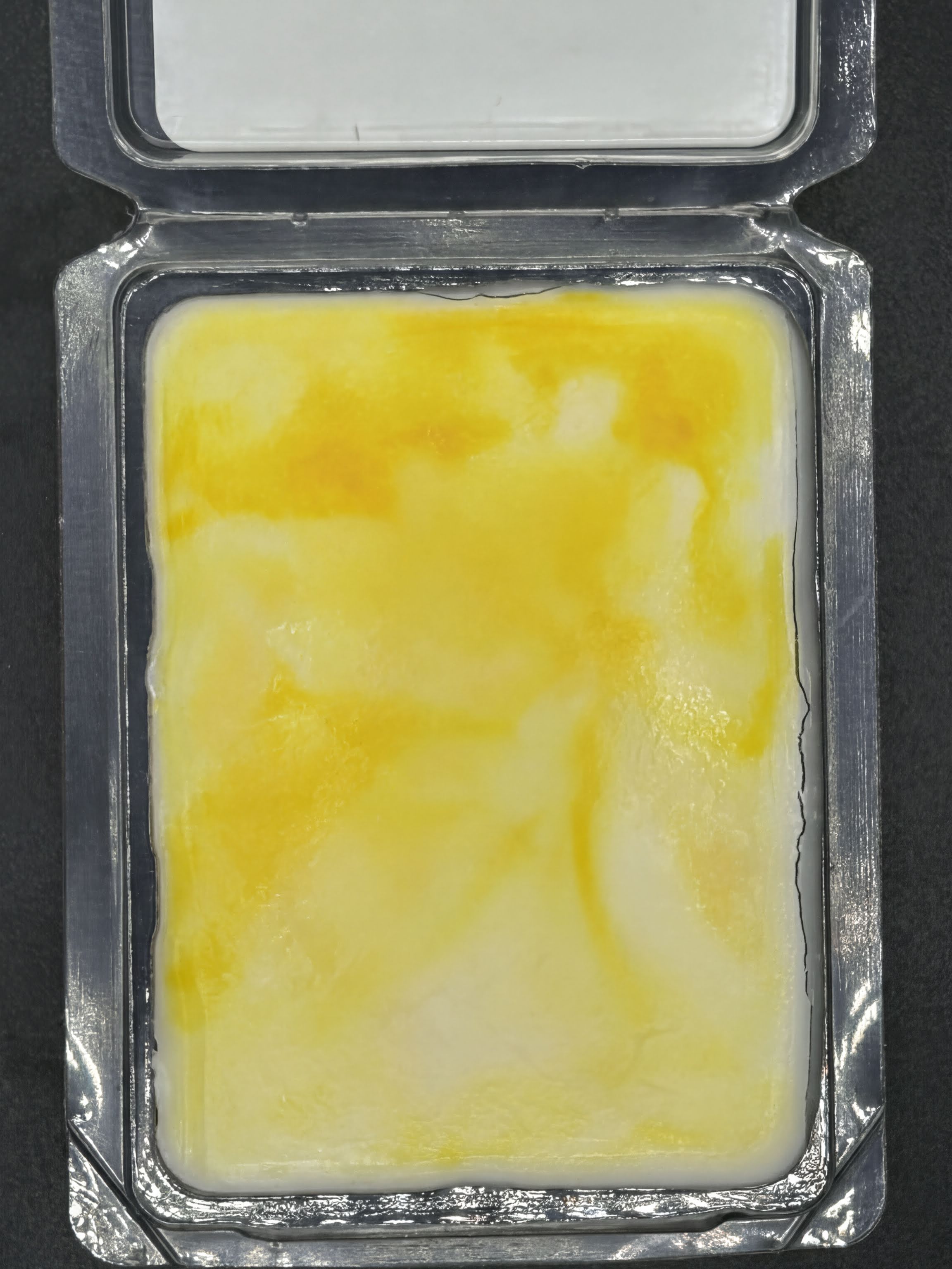 Yellow and white hand-poured wax melt in clear plastic clamshell packaging on dark background.