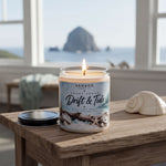 A lit candle in a jar sits on a wooden table, with a seashell and a black lid nearby. The candle is labeled "Sanbox Candle Co. Drift & Tide" and is set against a scenic view of the ocean and a rocky island in the distance.
