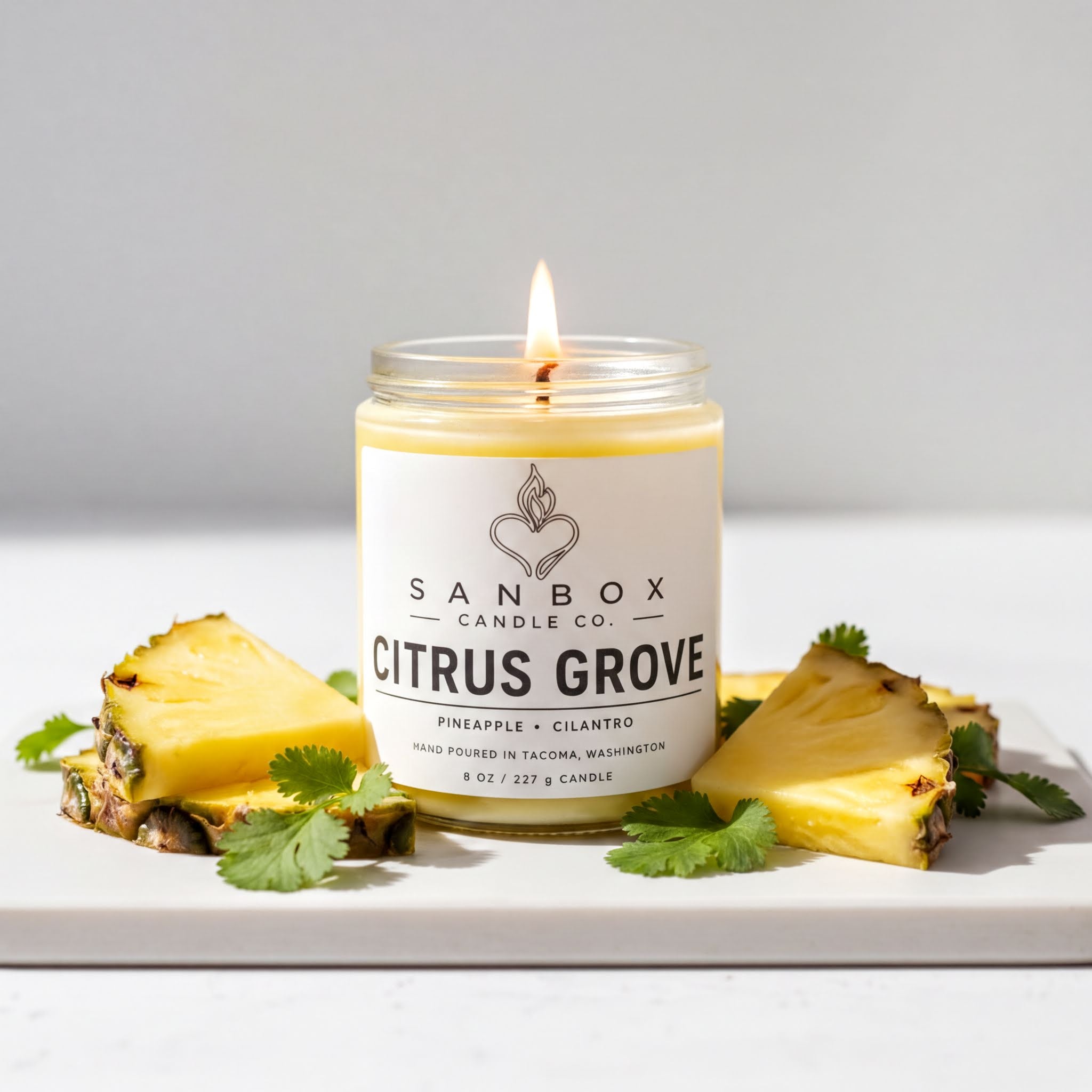 A lit candle in a jar, surrounded by pineapple slices and cilantro leaves, with a label that reads "SANBOX CANDLE CO. CITRUS GROVE Pineapple + Cilantro Hand Poured in Tacoma, Washington 8 oz / 227 swt"