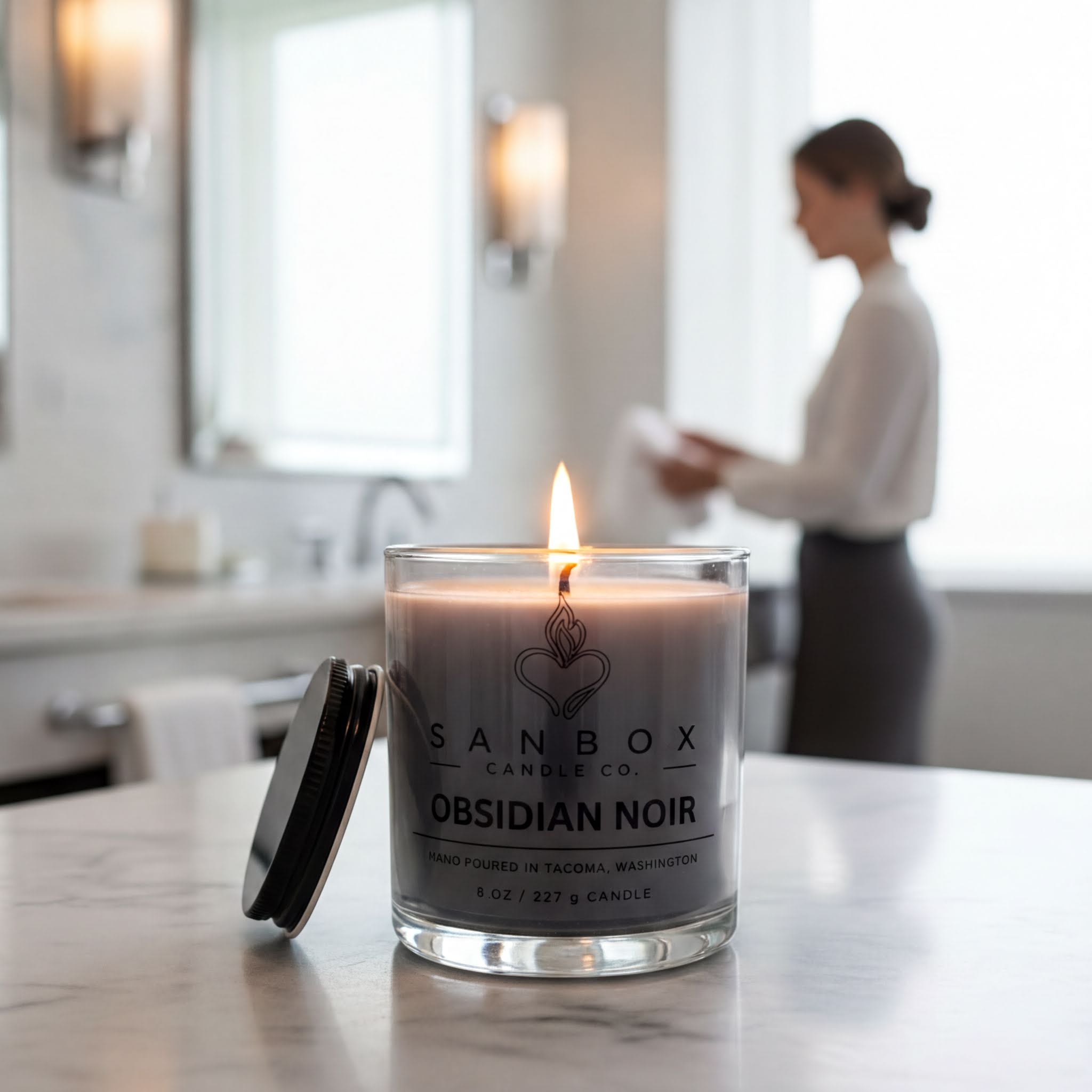 A lit candle in a glass container with a black lid, placed on a marble countertop. In the background, there is a blurred image of a person, likely a woman, standing near a window.