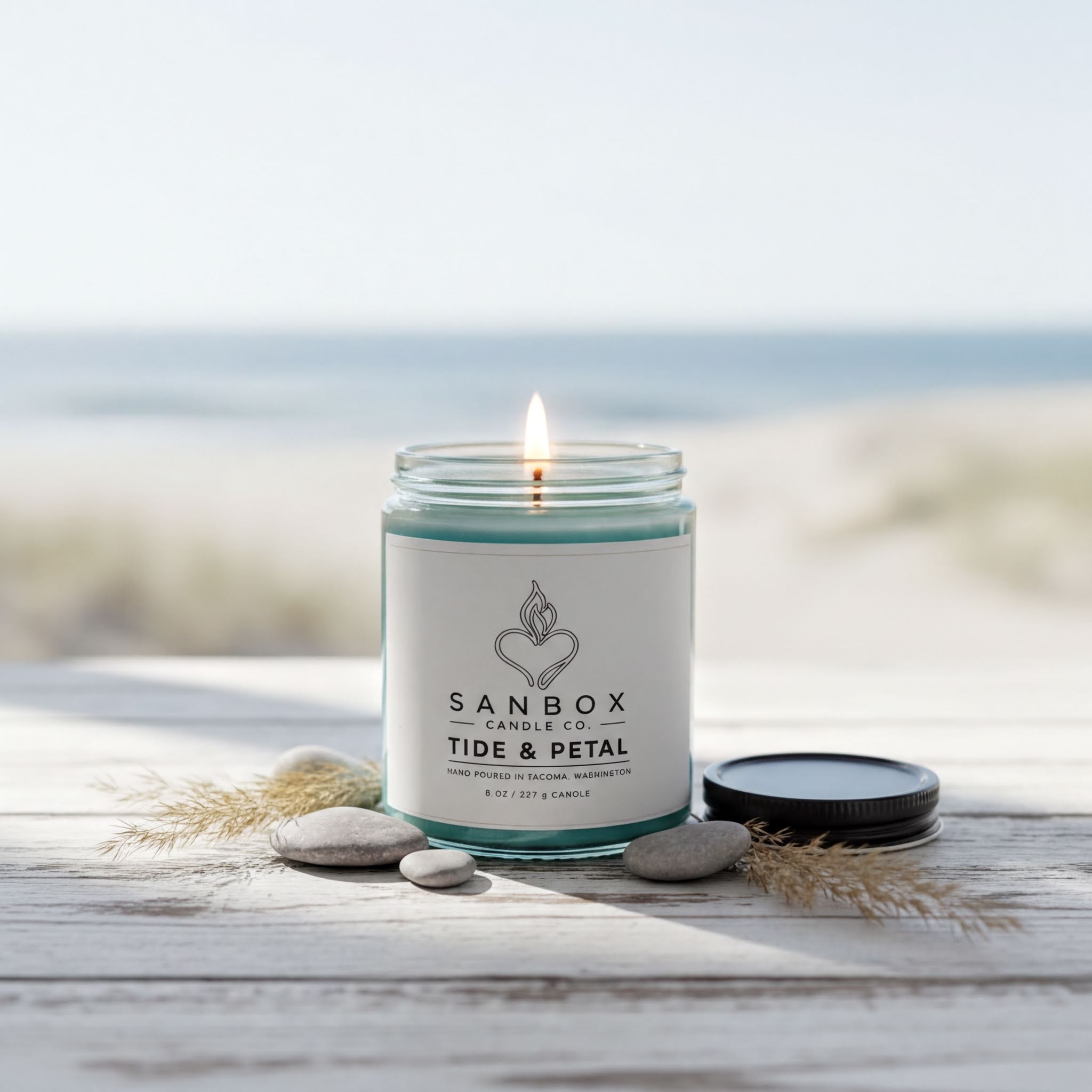 A lit candle in a glass jar with a label, surrounded by rocks and a blurred beach scene in the background.
