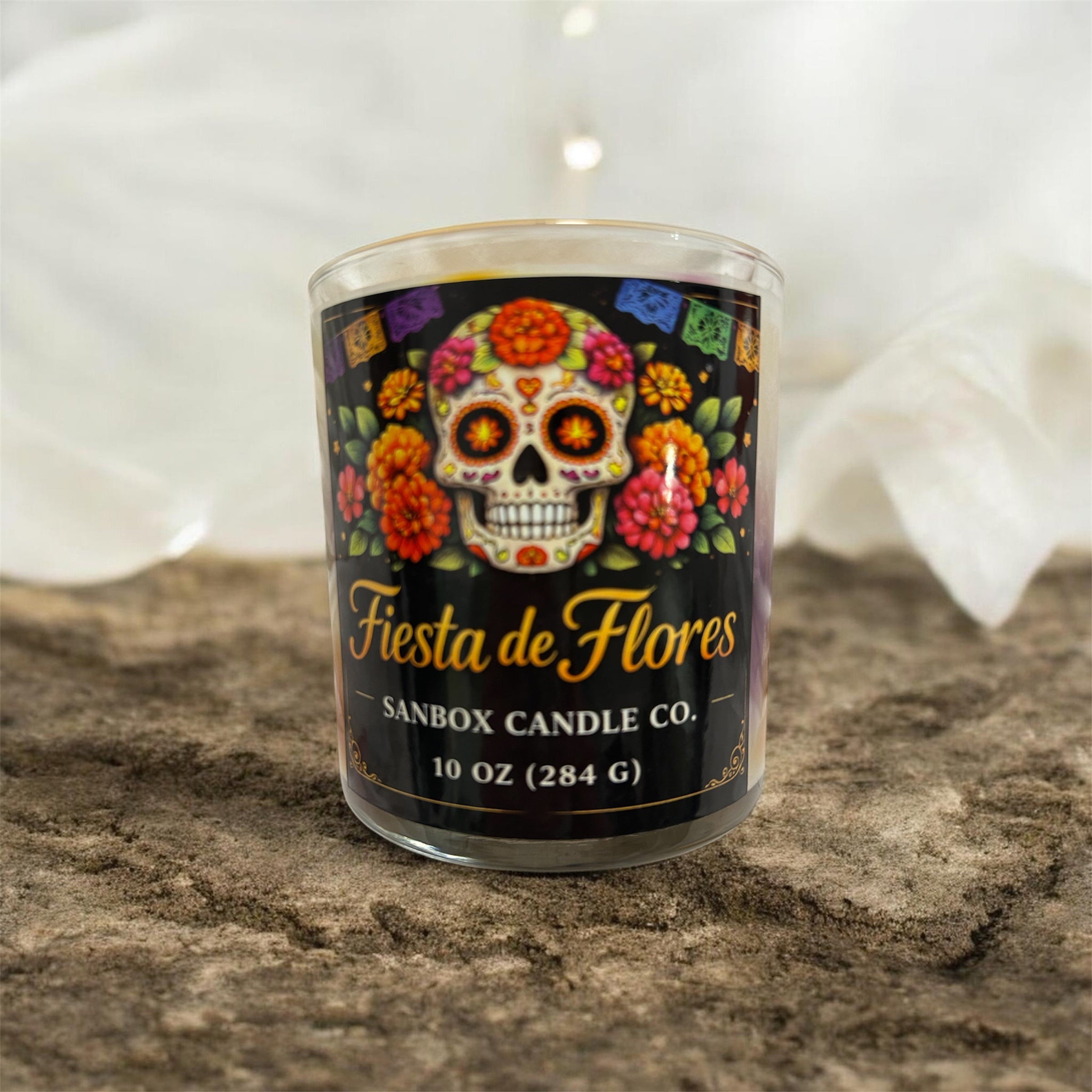 A glass candle holder with a label that reads "Fiesta de Flores" and "SANBOX CANDLE CO." The candle holder is placed on a surface with a textured background.