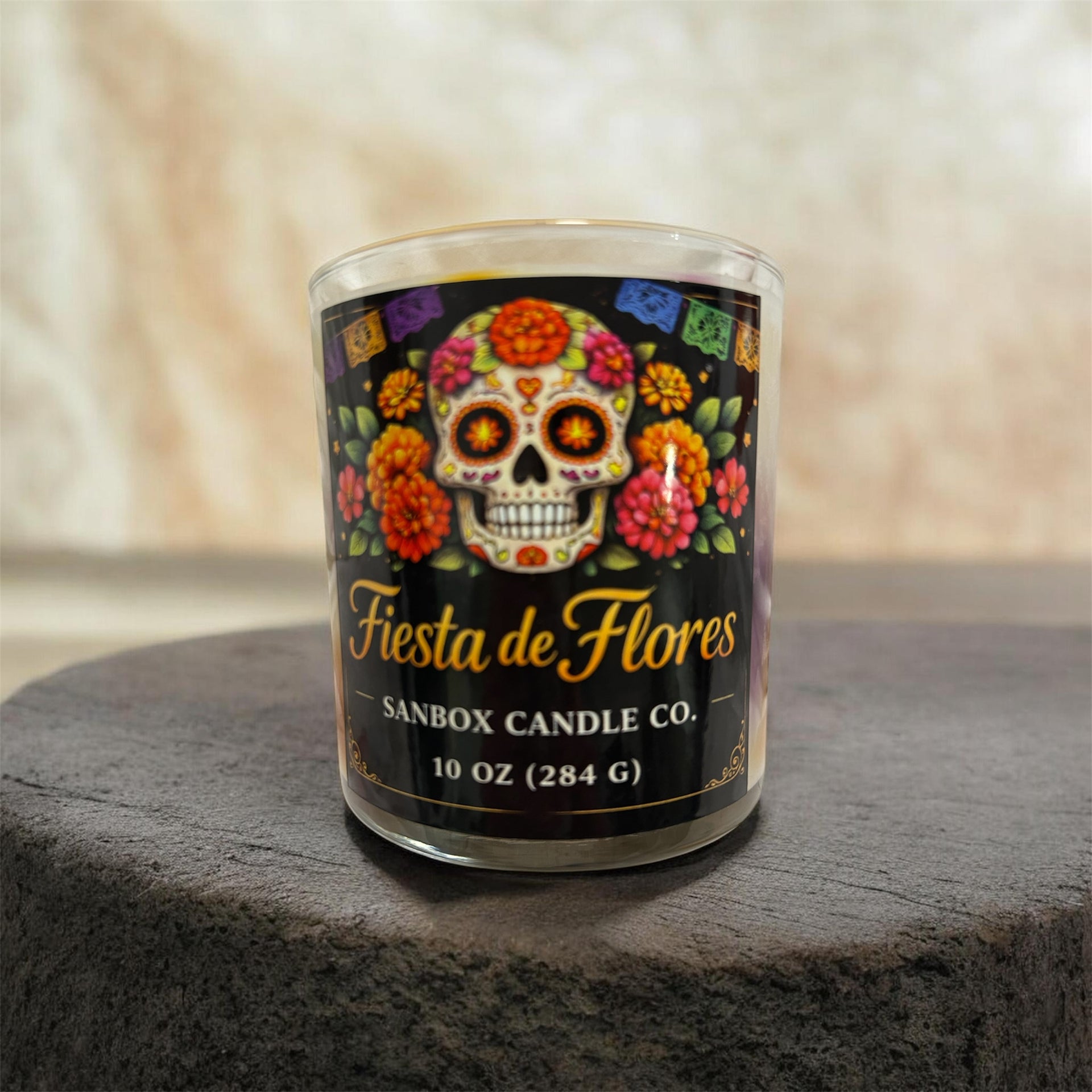 A glass candle holder with a label that reads "Fiesta de Flores" and "SANBOX CANDLE CO." The candle holder is placed on a wooden surface.
