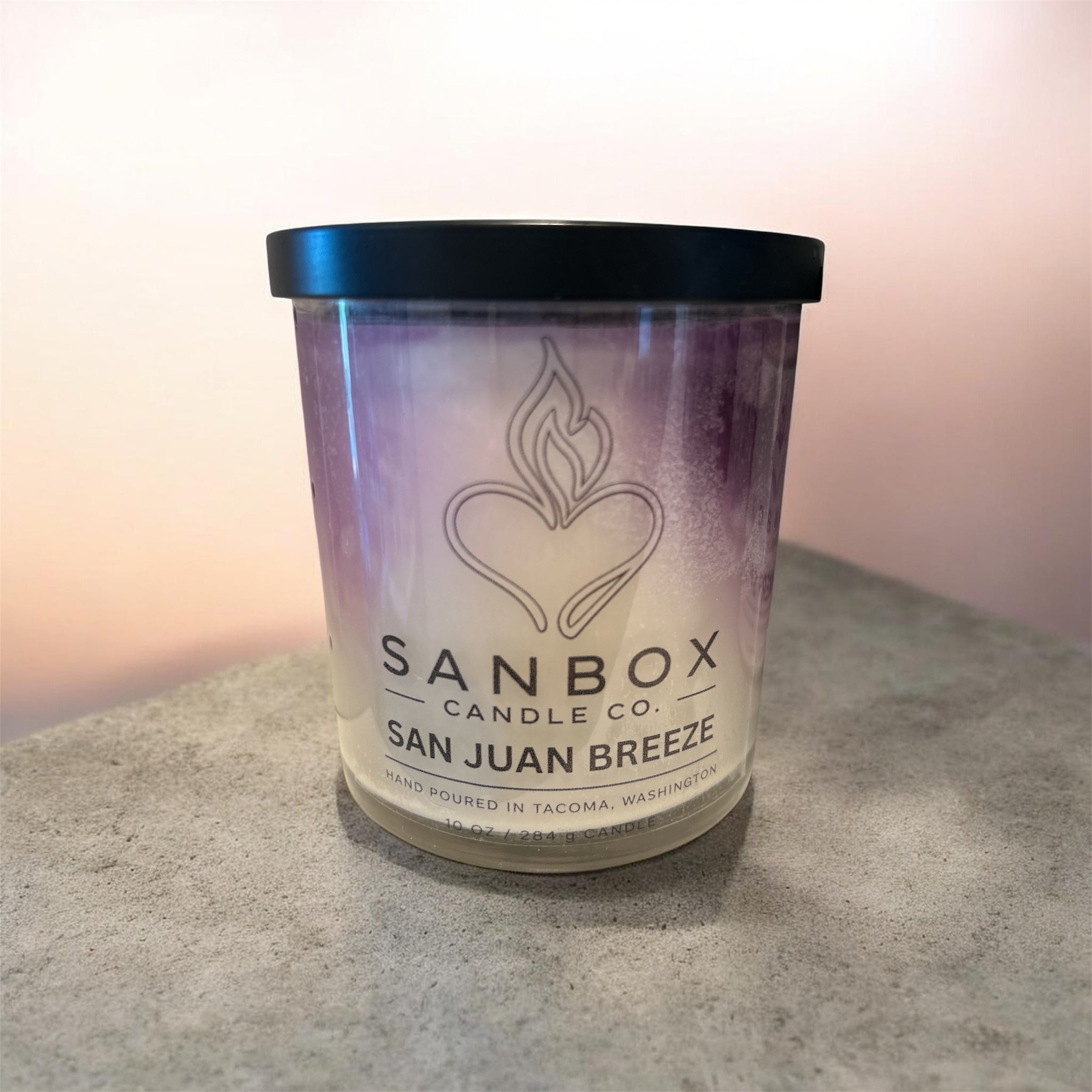 A candle in a clear glass jar with a black lid, featuring a label with text and a graphic design.